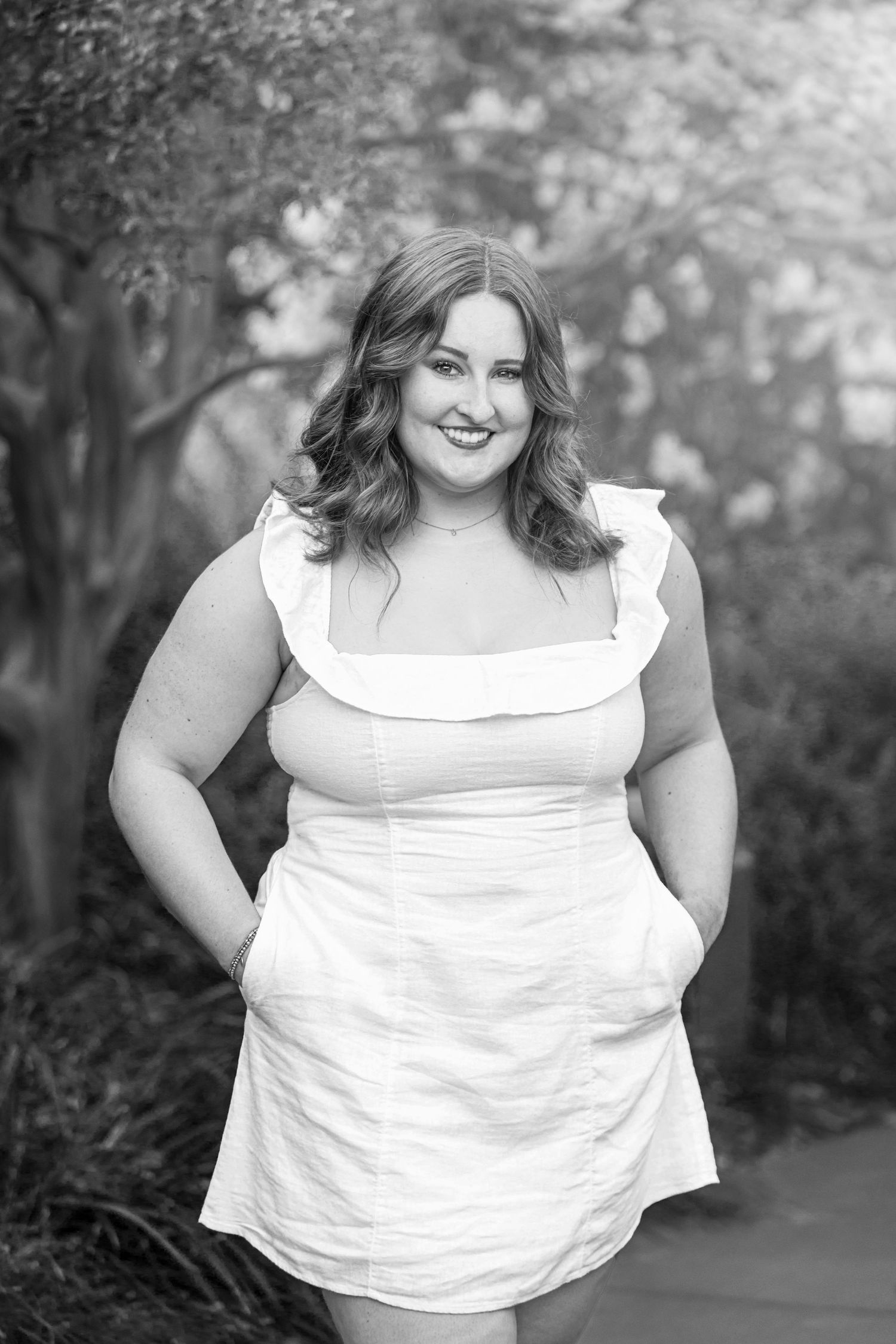 Confident woman in white ruffled sundress poses outdoors among blooming trees in black and white photo.