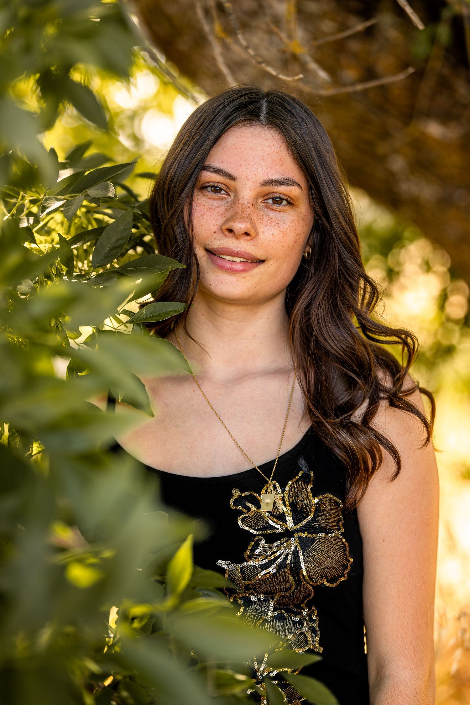 Young woman with freckles and brown wavy hair smiling among green sunlit leaves wearing black top.