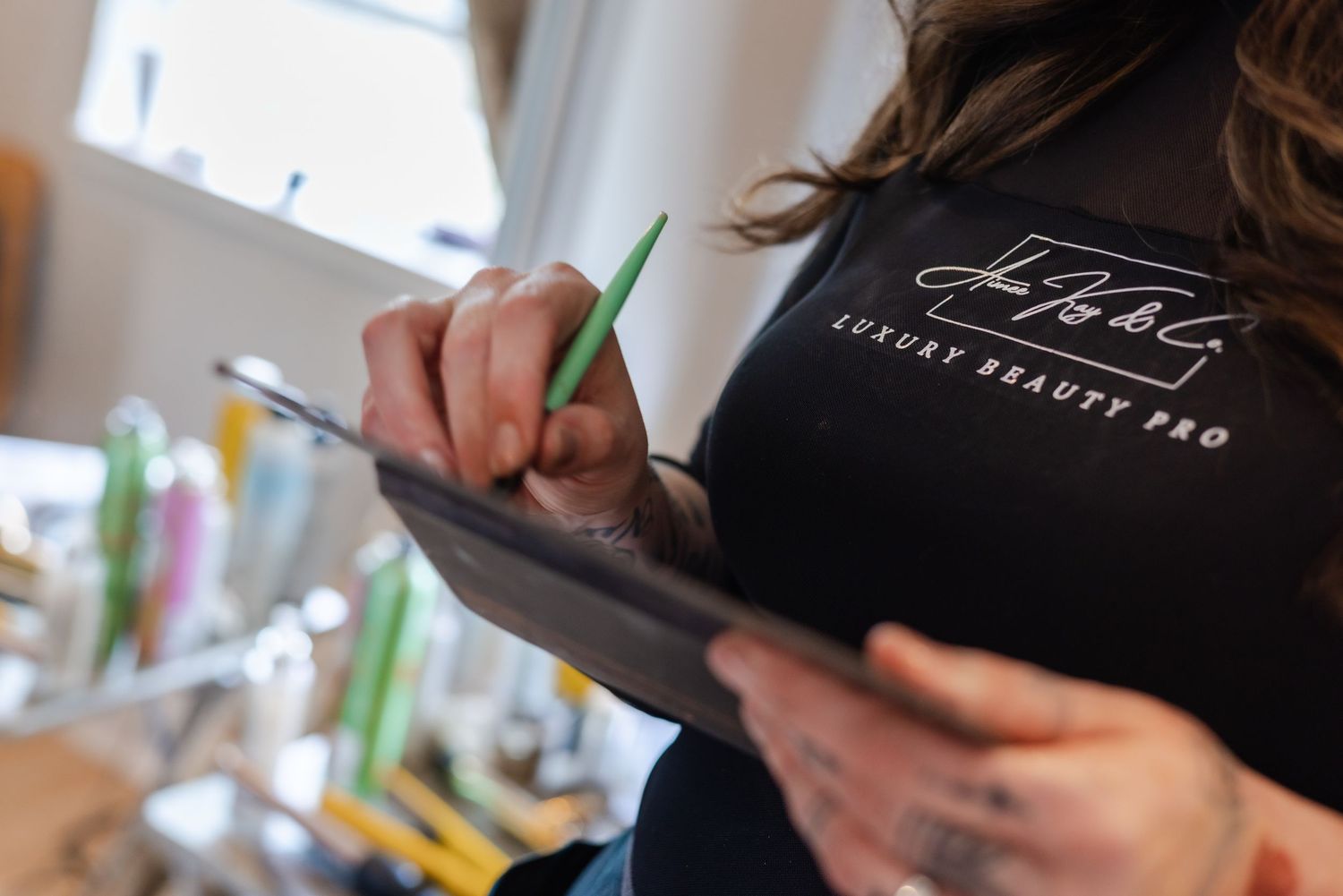 Beauty professional in black Luxury Beauty Pro shirt writing on clipboard with green pen.
