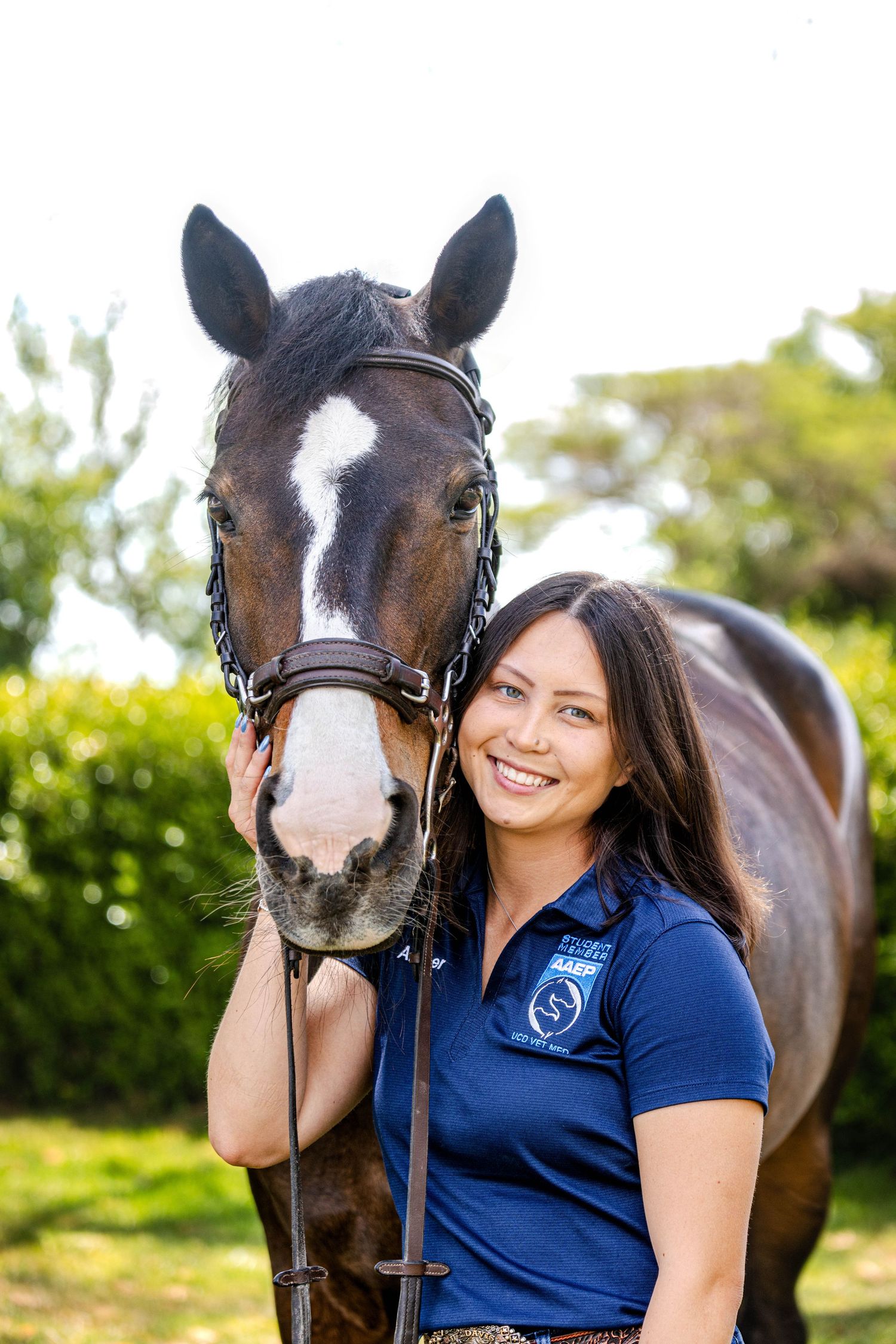 Smiling brunette woman in blue polo shirt poses with dark bay horse with white blaze.