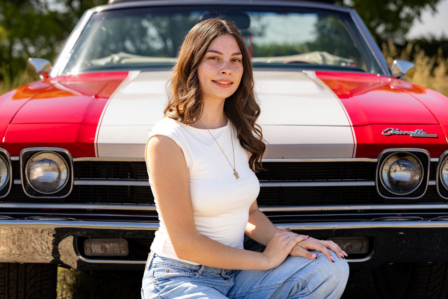 Young woman in white top and jeans poses in front of a classic red and white Chevrolet muscle car outdoors.