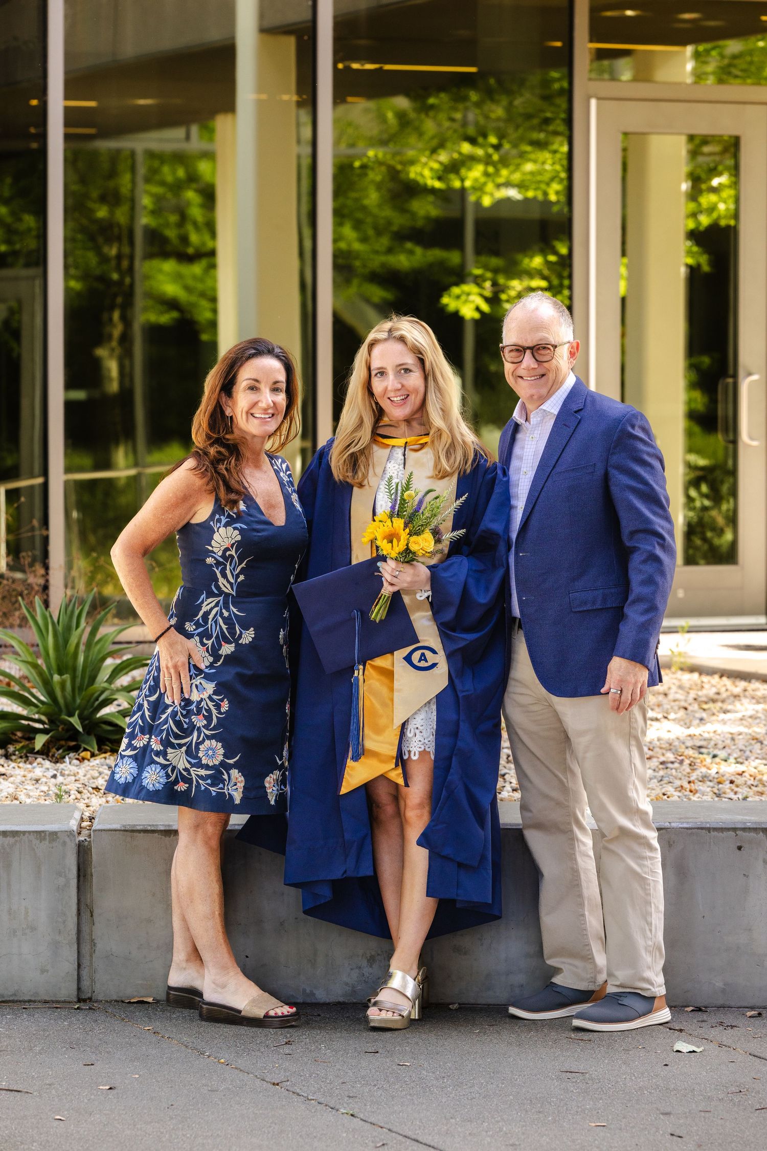 A graduate in blue cap and gown holds yellow flowers, posing with two proud family members outside a building.