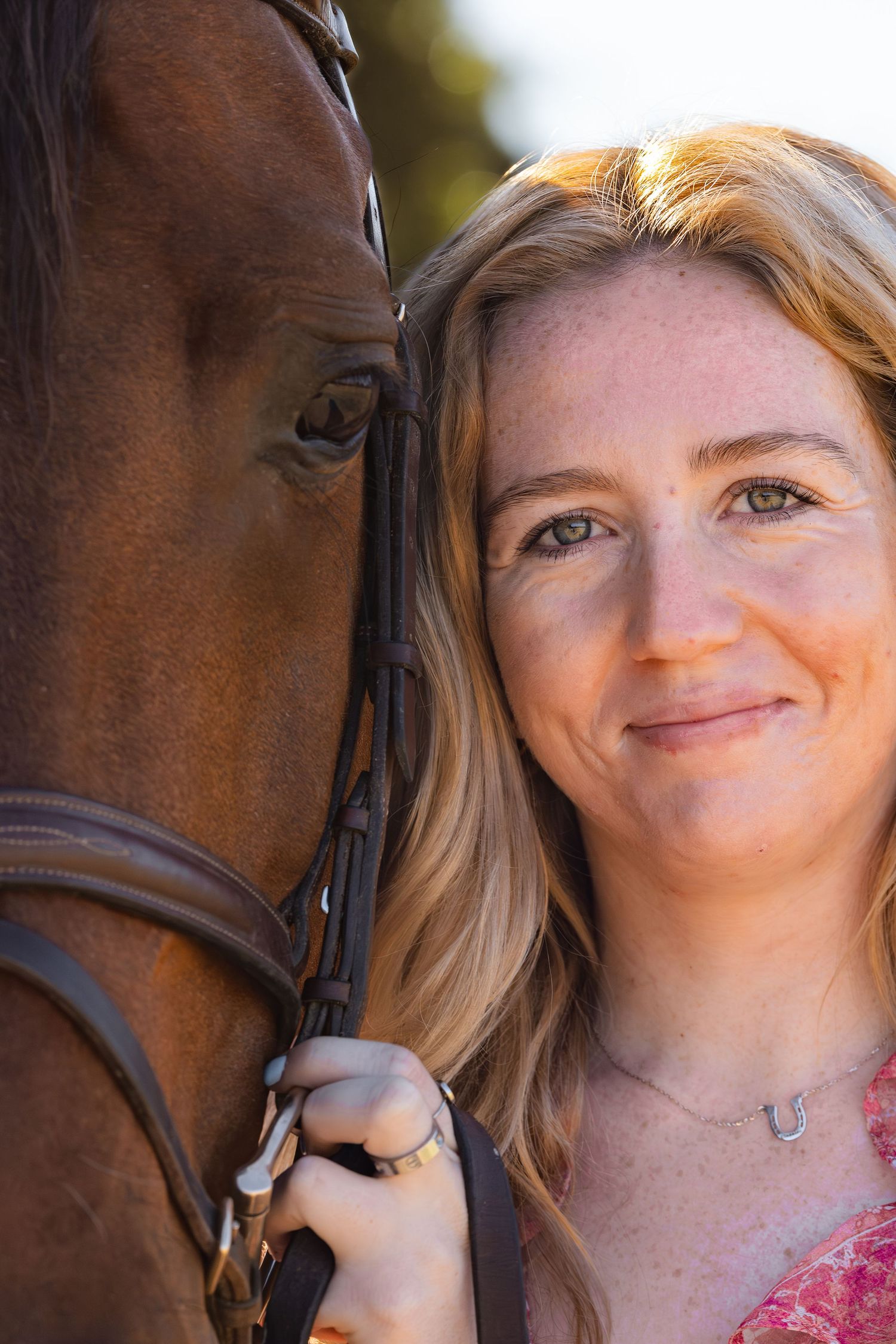 Close-up portrait of a smiling blonde woman with freckles posing cheek-to-cheek with a bay horse.