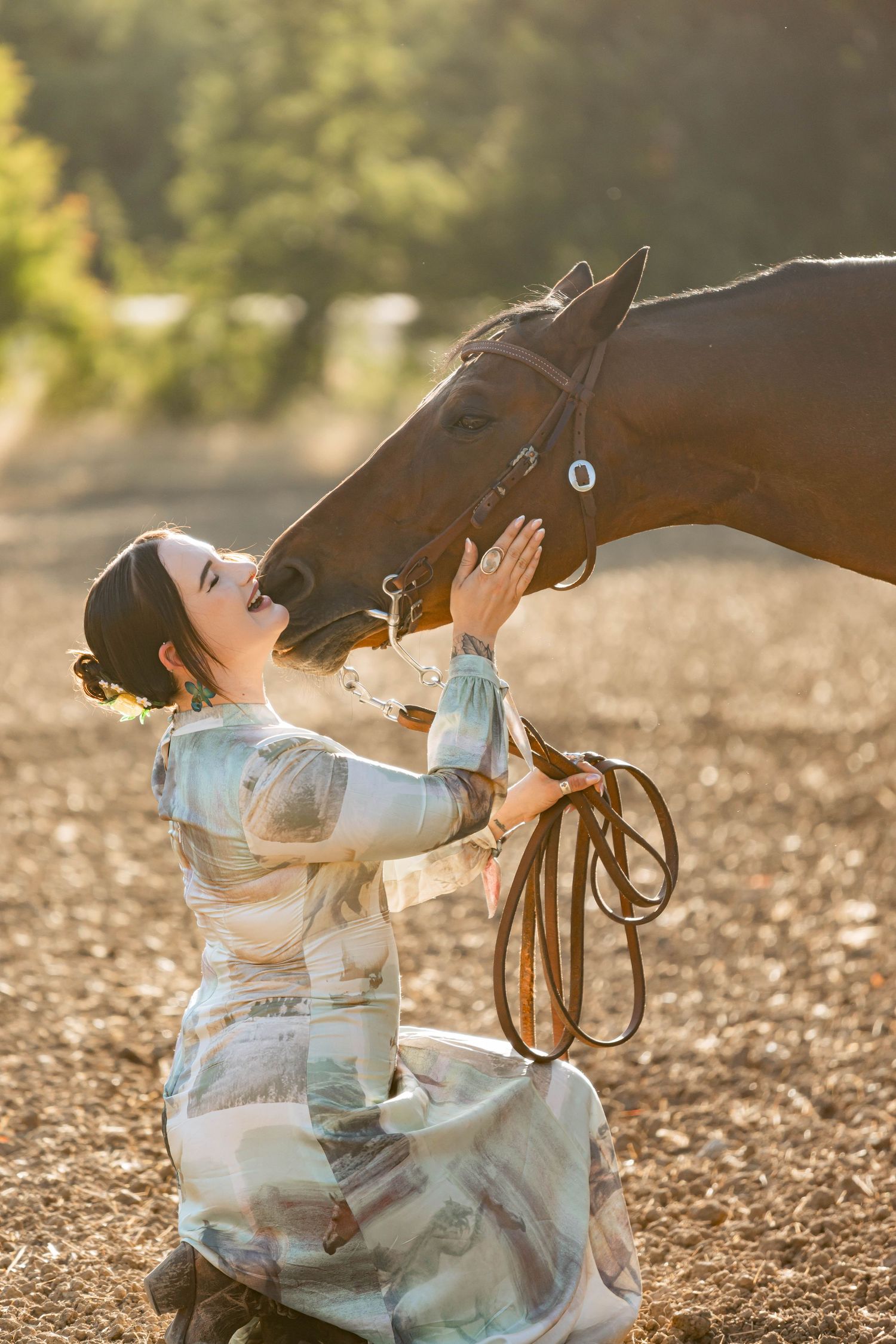 Woman in floral dress kneels laughing as a chestnut horse nuzzles her in warm light.