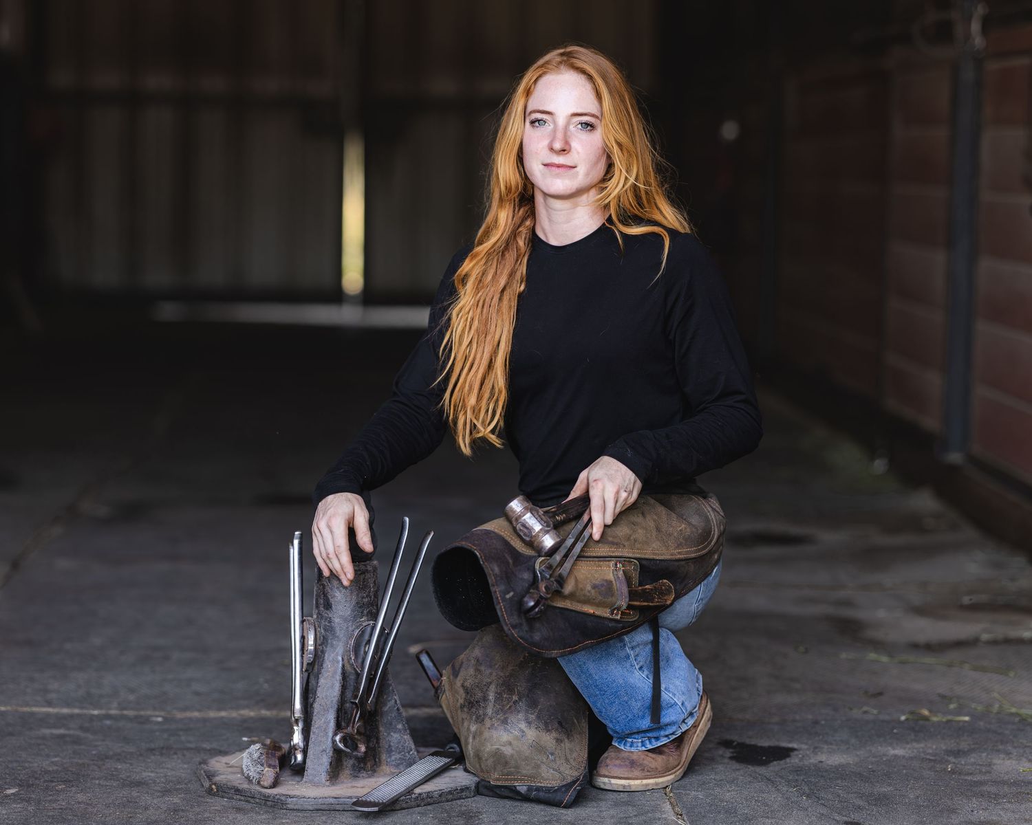 Young redhead woman kneeling in a barn with farrier tools and a horse saddle.