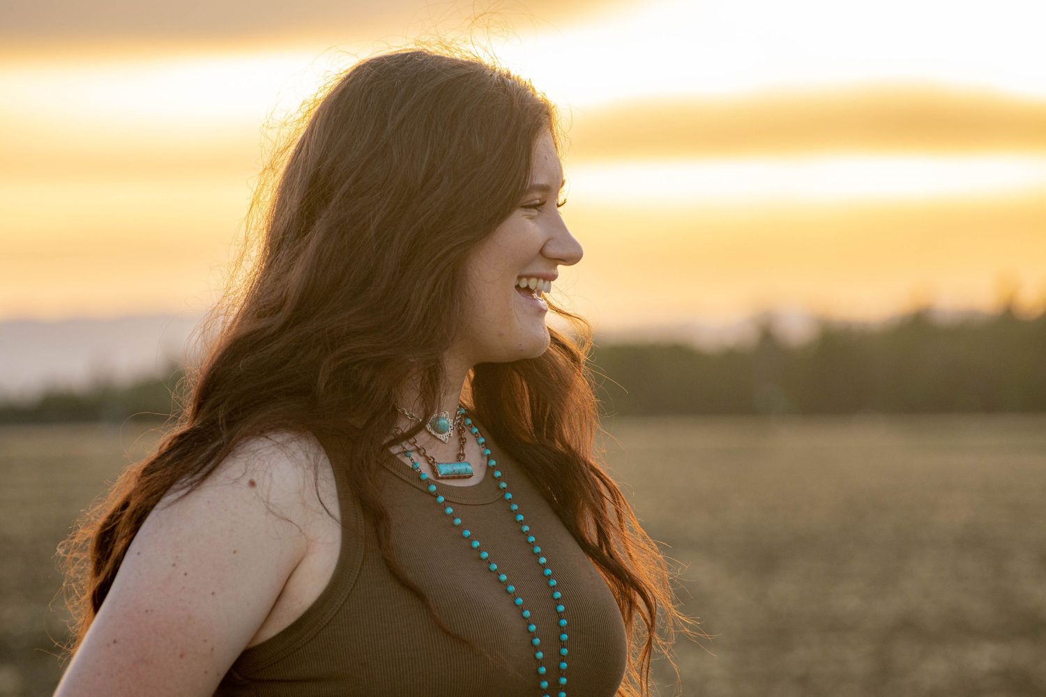 Young woman with long auburn hair smiles in profile against vibrant golden sunset sky.