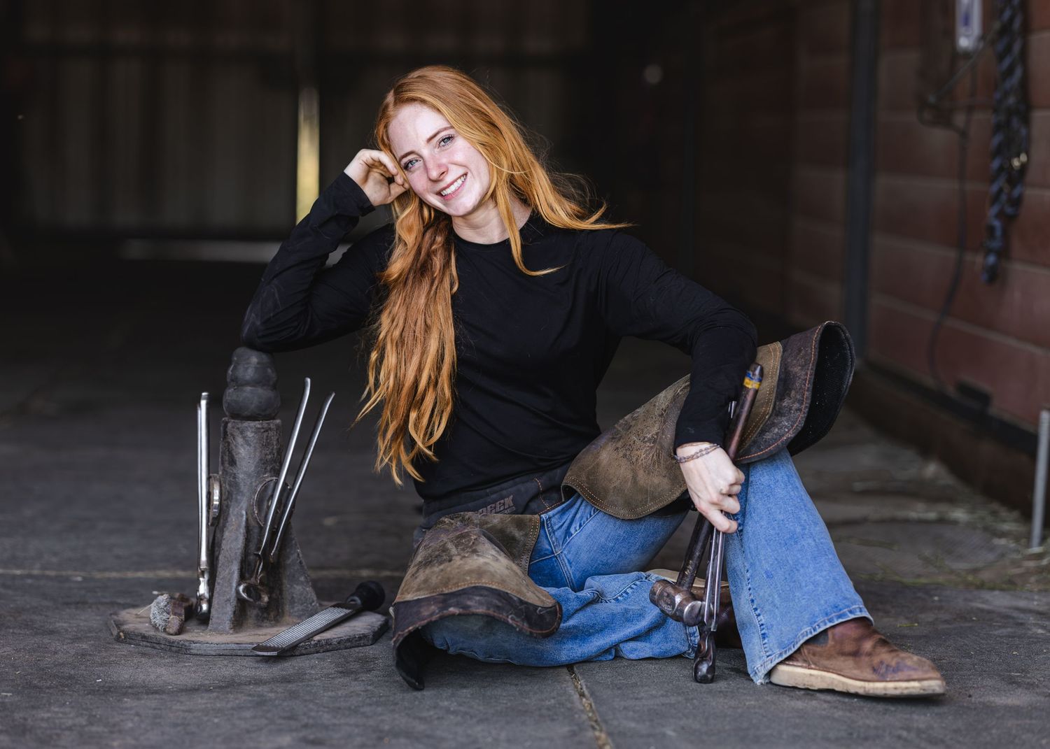 Smiling young woman with long red hair sitting on barn floor wearing black top, jeans, and cowboy boots.