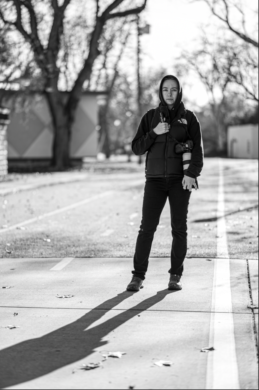 Black and white photo of a photographer standing on a road holding a camera in an urban outdoor setting.