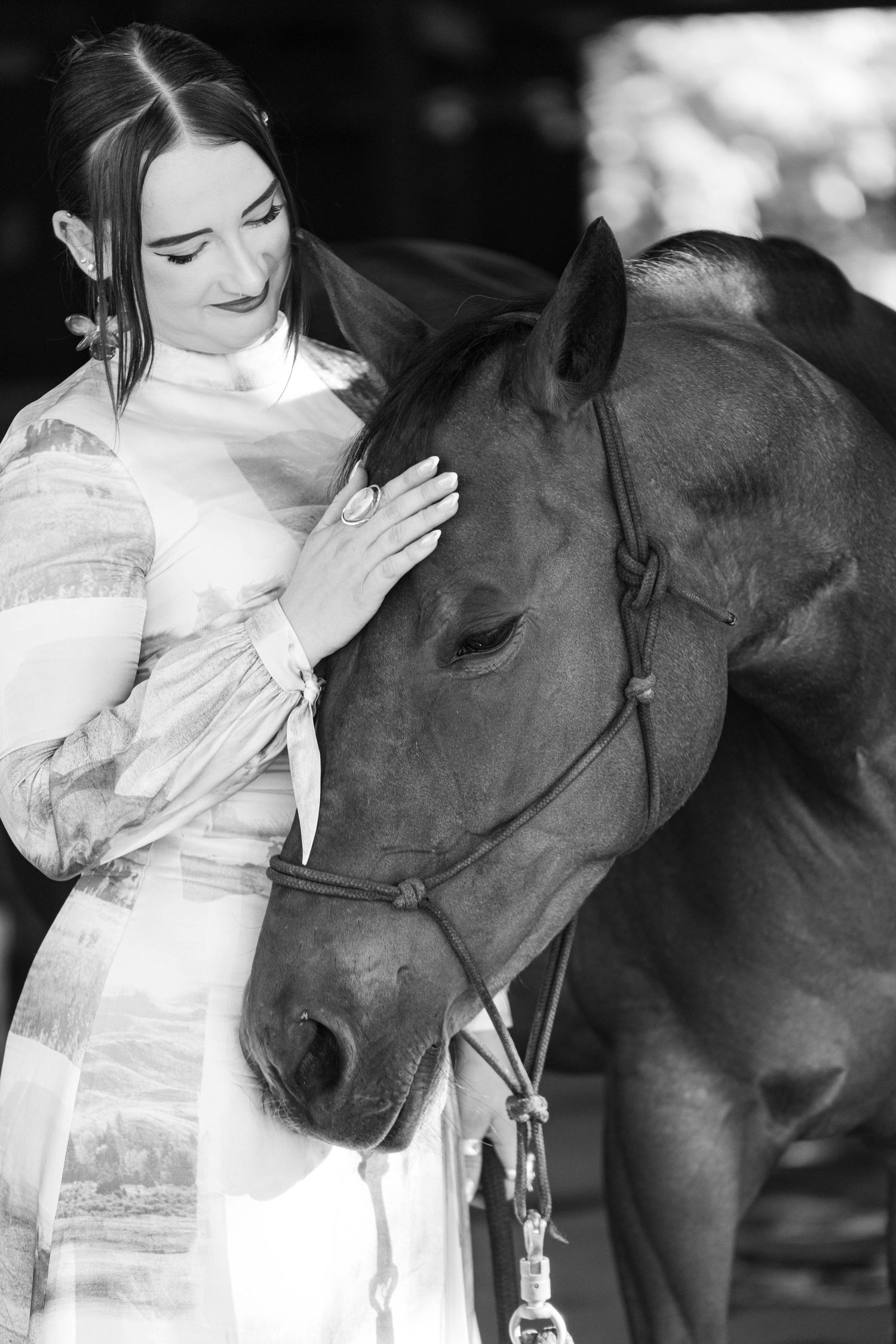 Black and white portrait of elegant woman in white dress tenderly touching a horse's face in stable setting.