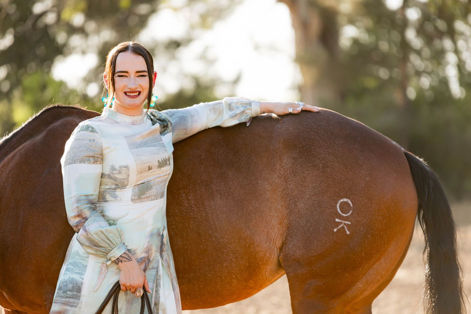 Smiling woman in floral dress stands beside a chestnut horse in soft natural light.