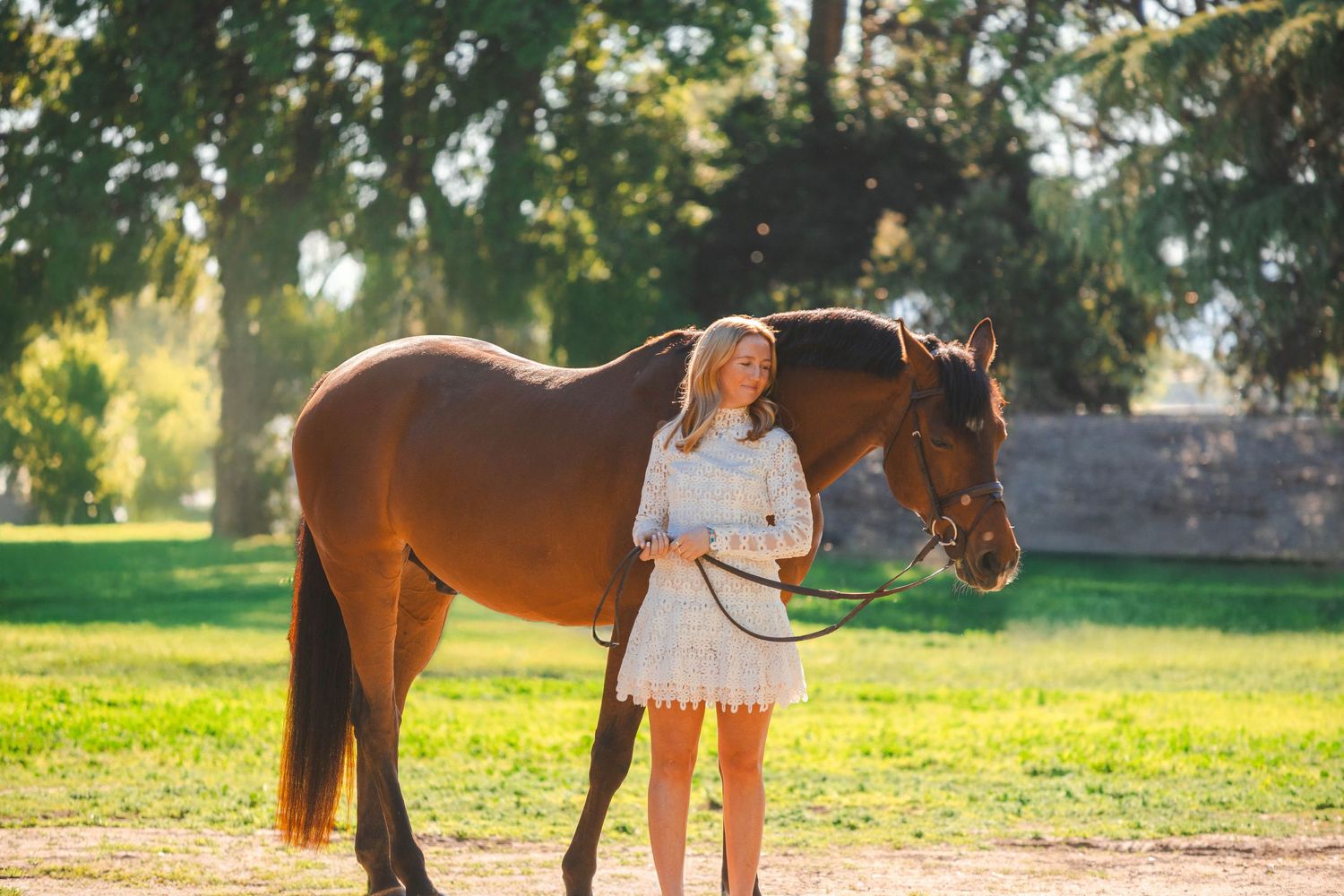 Young blonde woman in white dress stands with a chestnut horse in sunny green field.