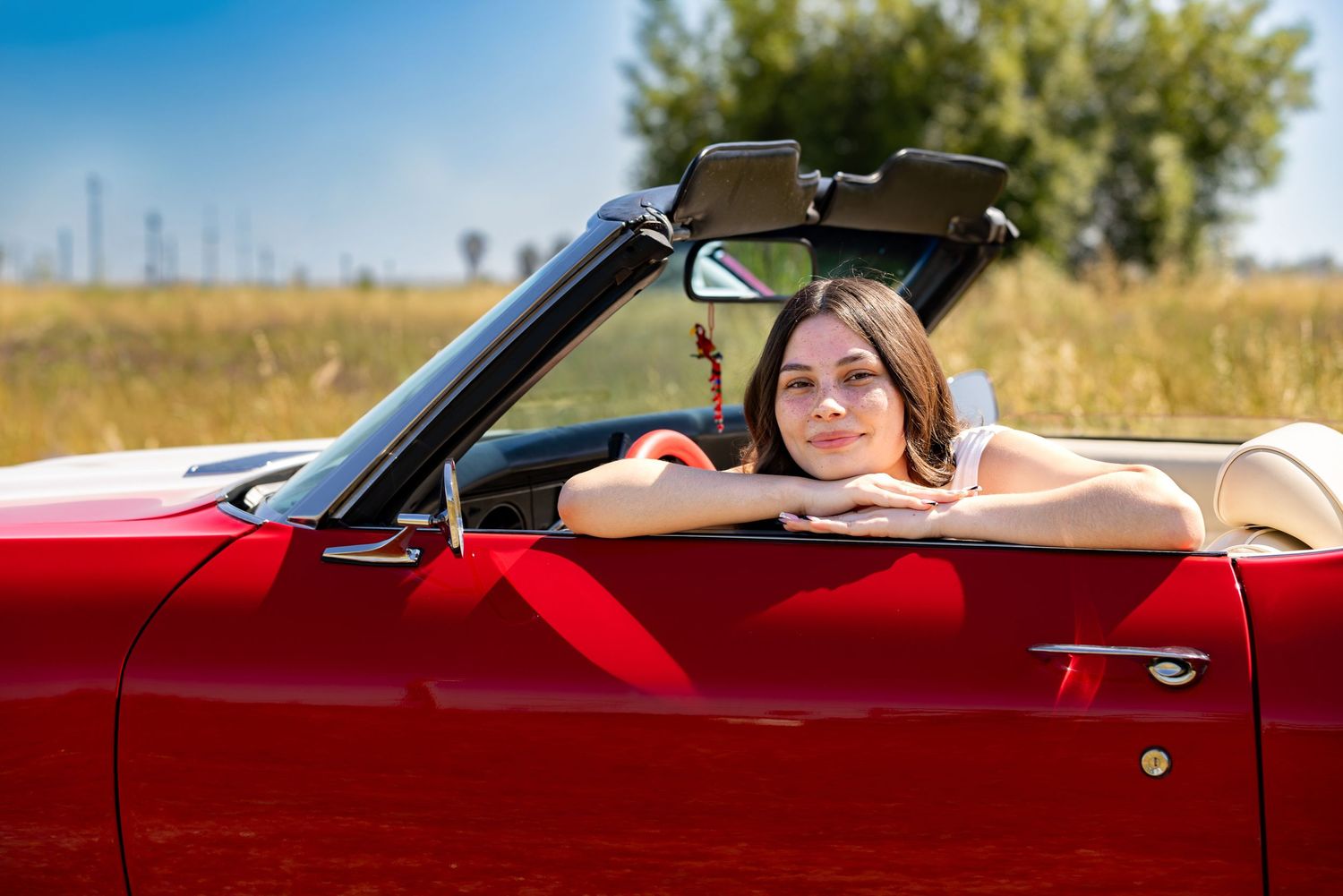 Young woman smiling in a red convertible car on a sunny day in a field.