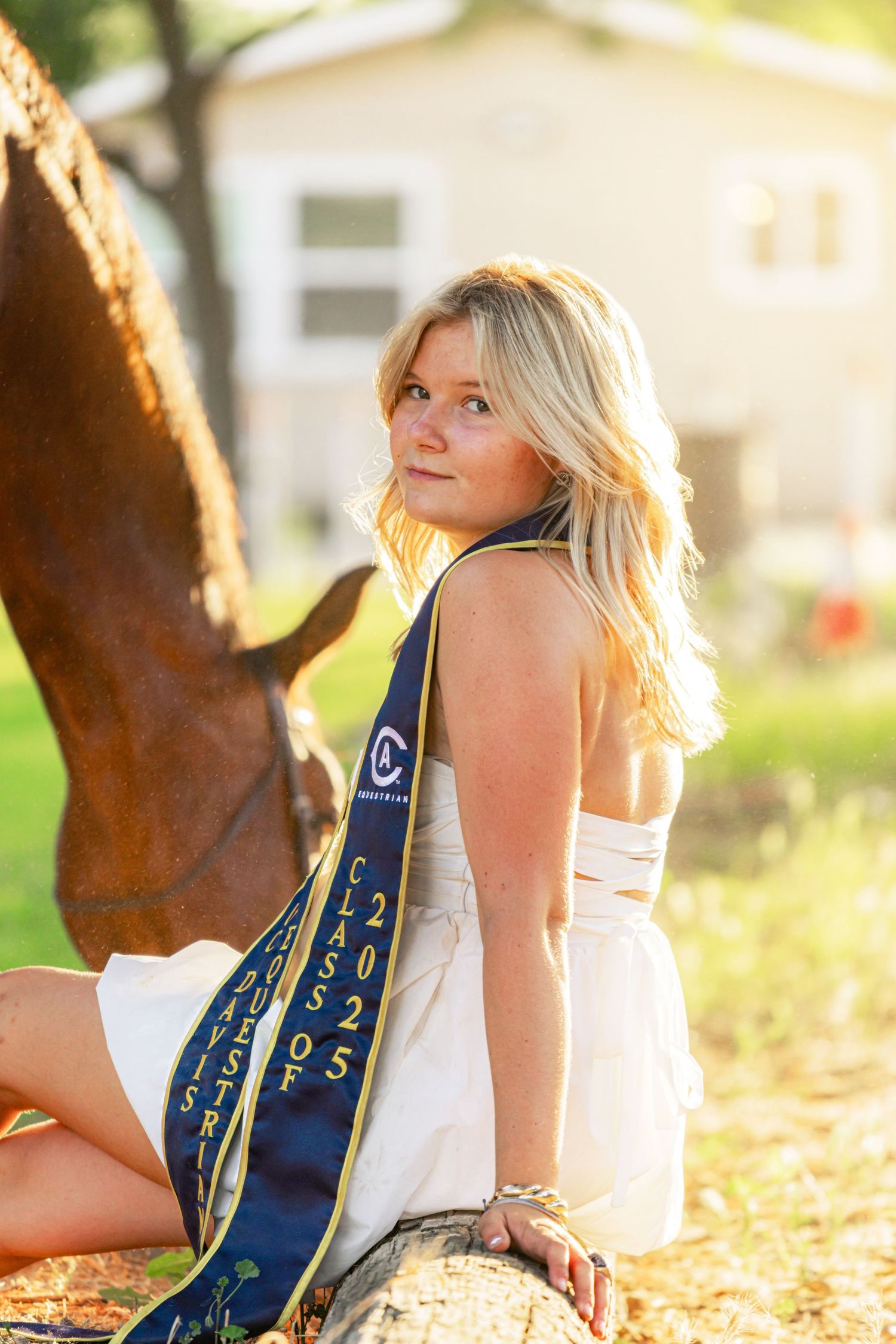 Blonde teen in white dress wearing Class of 2025 graduation sash seated near horse in golden sunlight.