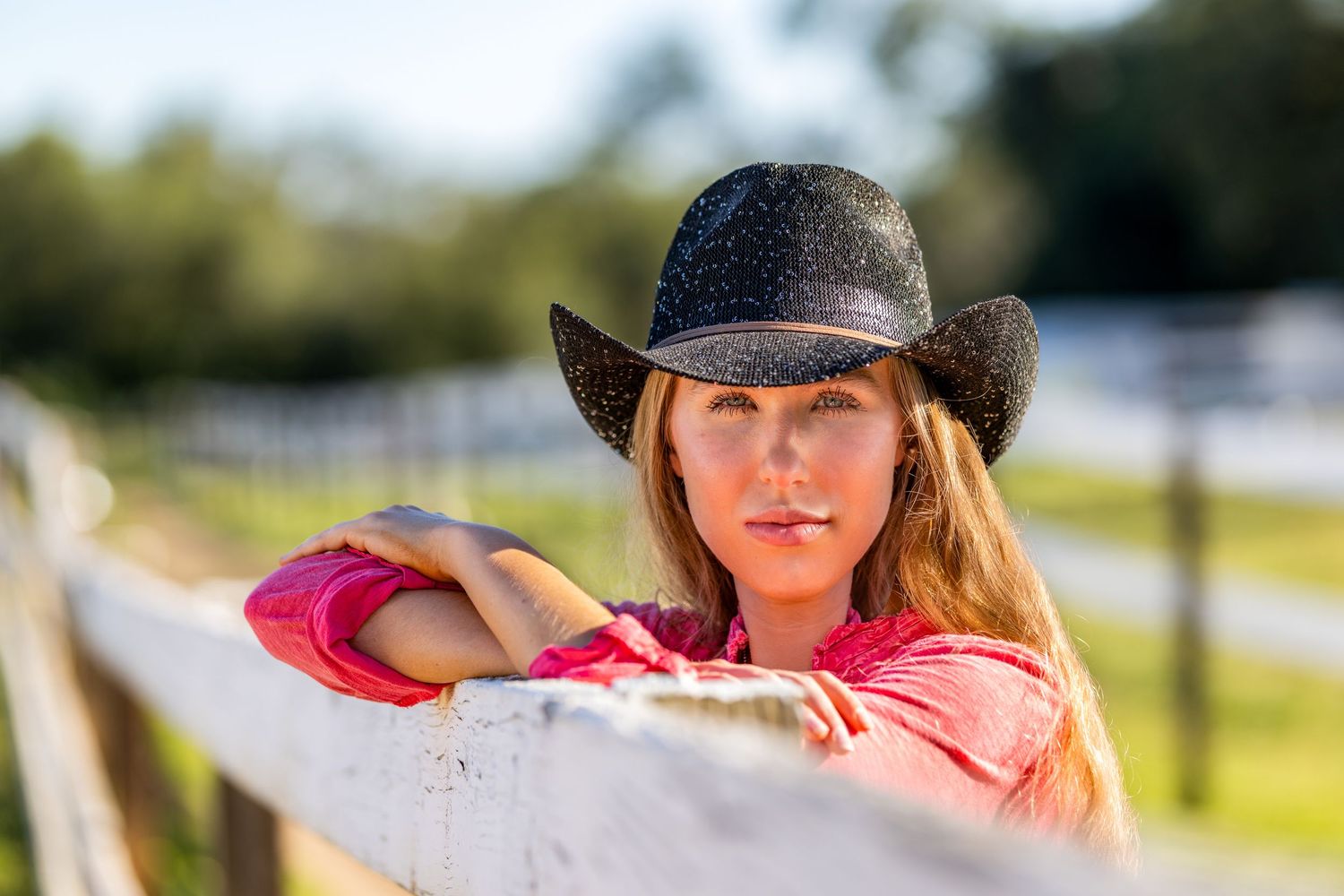 Woman in pink shirt and black cowboy hat resting on white wooden fence in golden light.