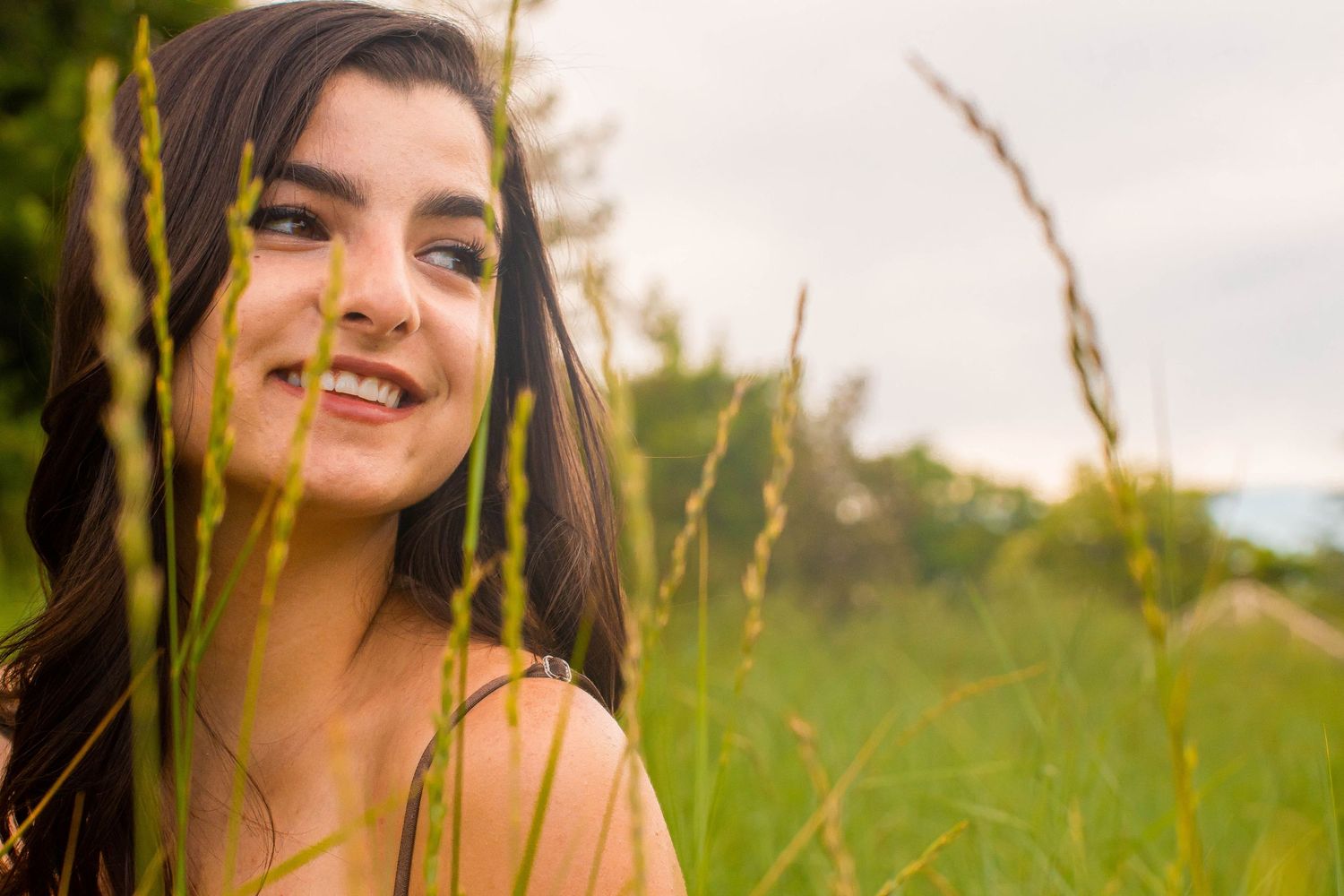 Smiling young woman with dark hair posing among tall green grass in a field on a cloudy day.