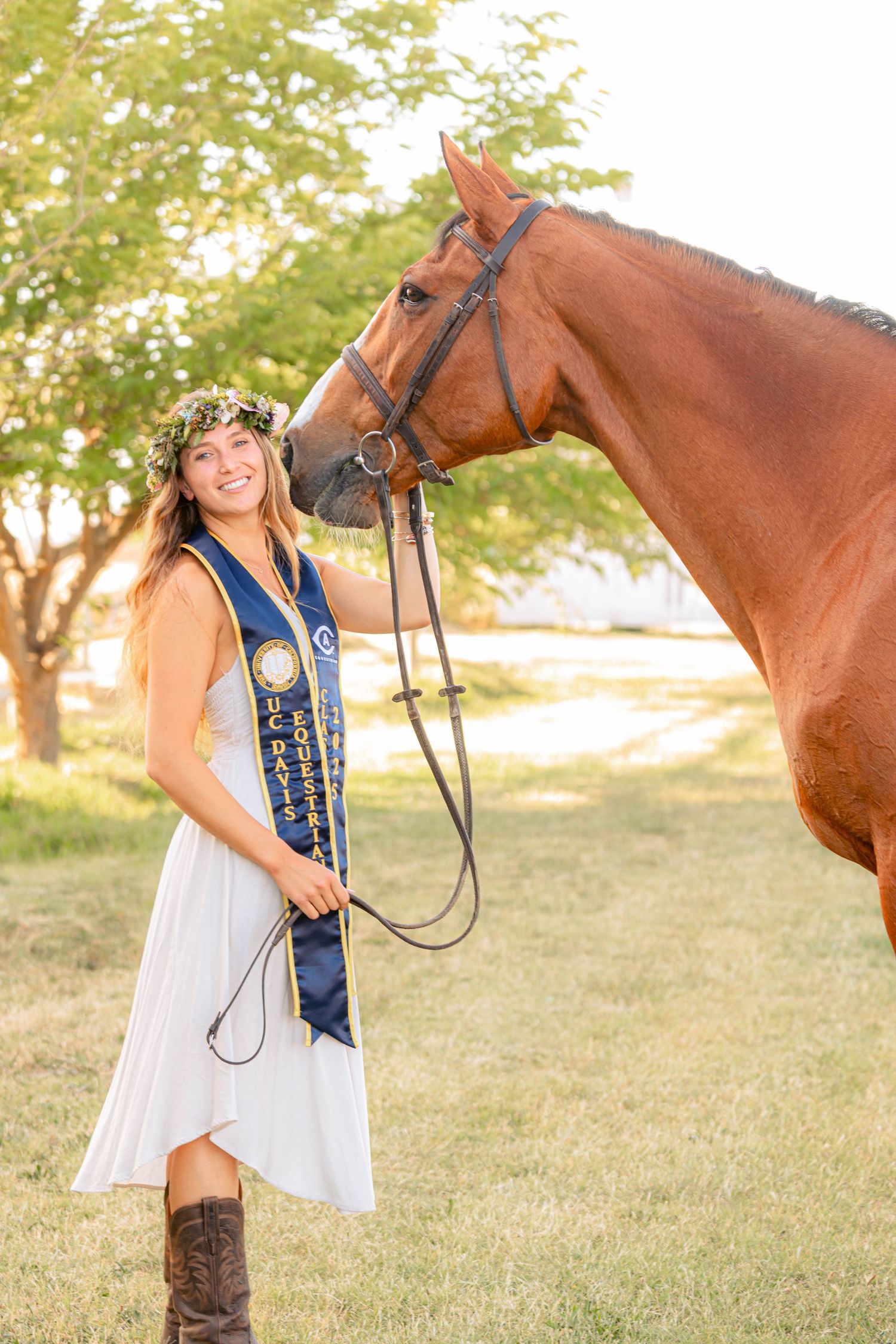 Smiling graduate in white dress and blue sash poses with chestnut horse outdoors, wearing floral crown and cowboy boots.