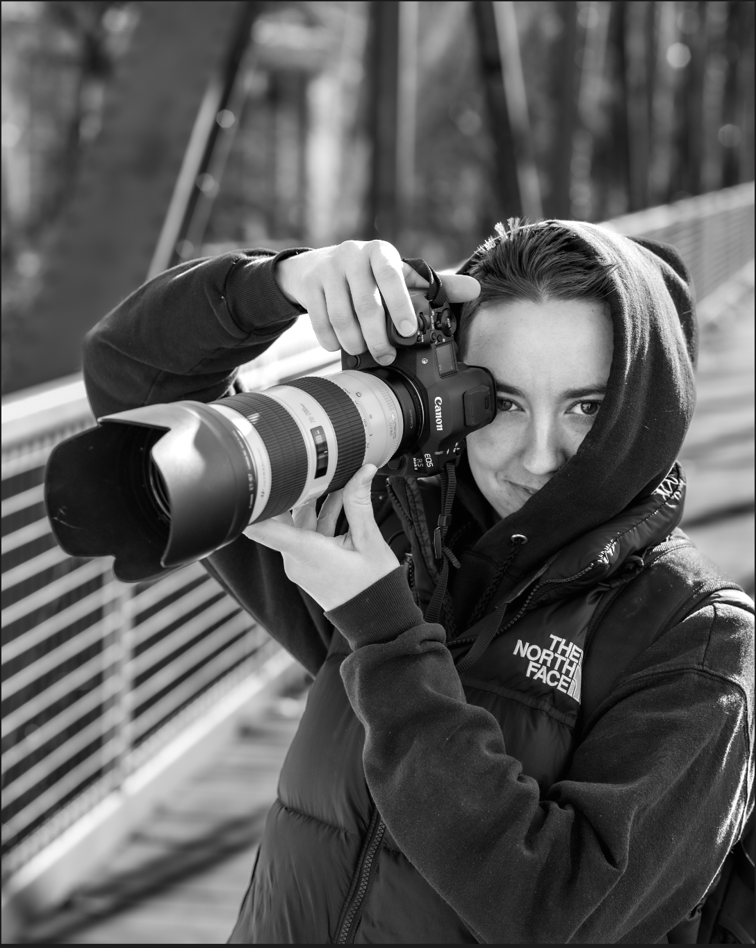 The photographer, Mattie, in a North Face hoodie holding a Canon camera with telephoto lens in black and white.