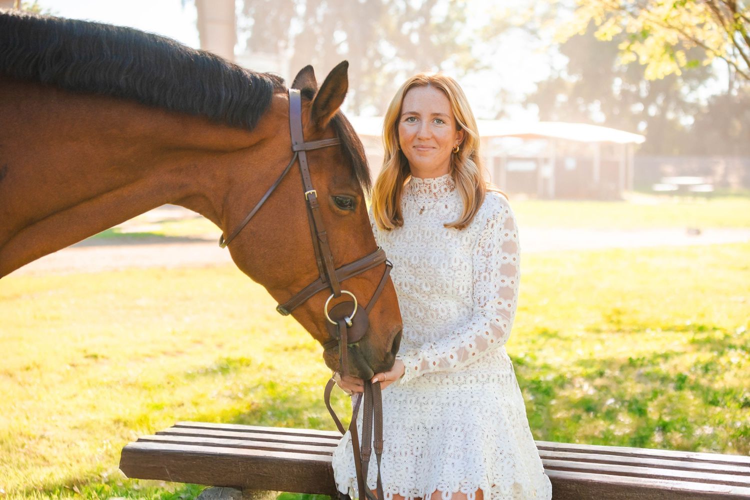 Blonde woman in white lace dress standing beside a chestnut horse in sunny park.