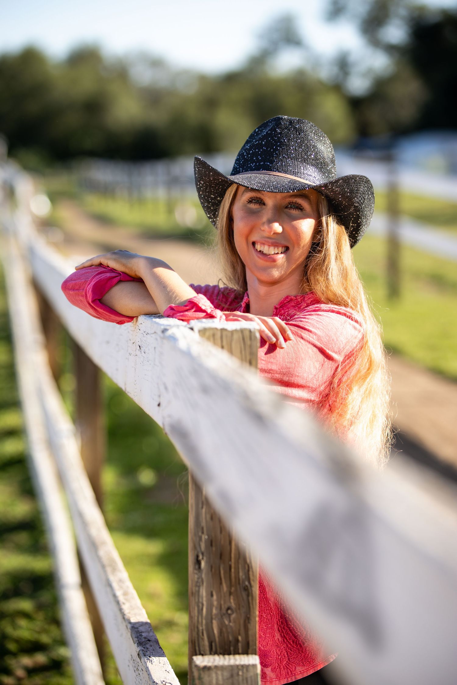 Smiling blonde woman in a black cowboy hat and pink shirt leaning on a white wooden fence at a sunny ranch.