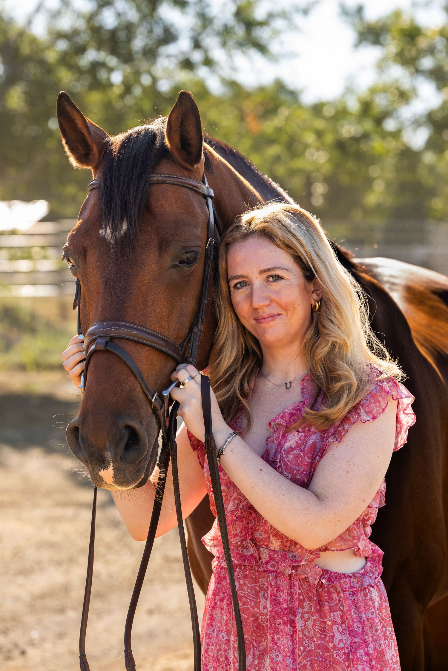 Woman in pink floral dress posing with a bay horse at a sunny equestrian farm.