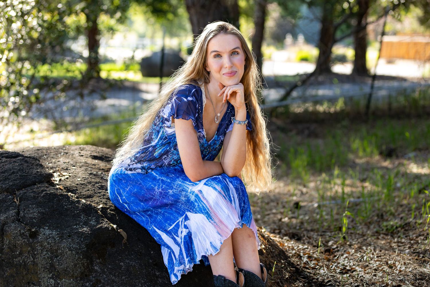 Young blonde woman in blue floral dress sitting on rock in sunlit wooded setting.