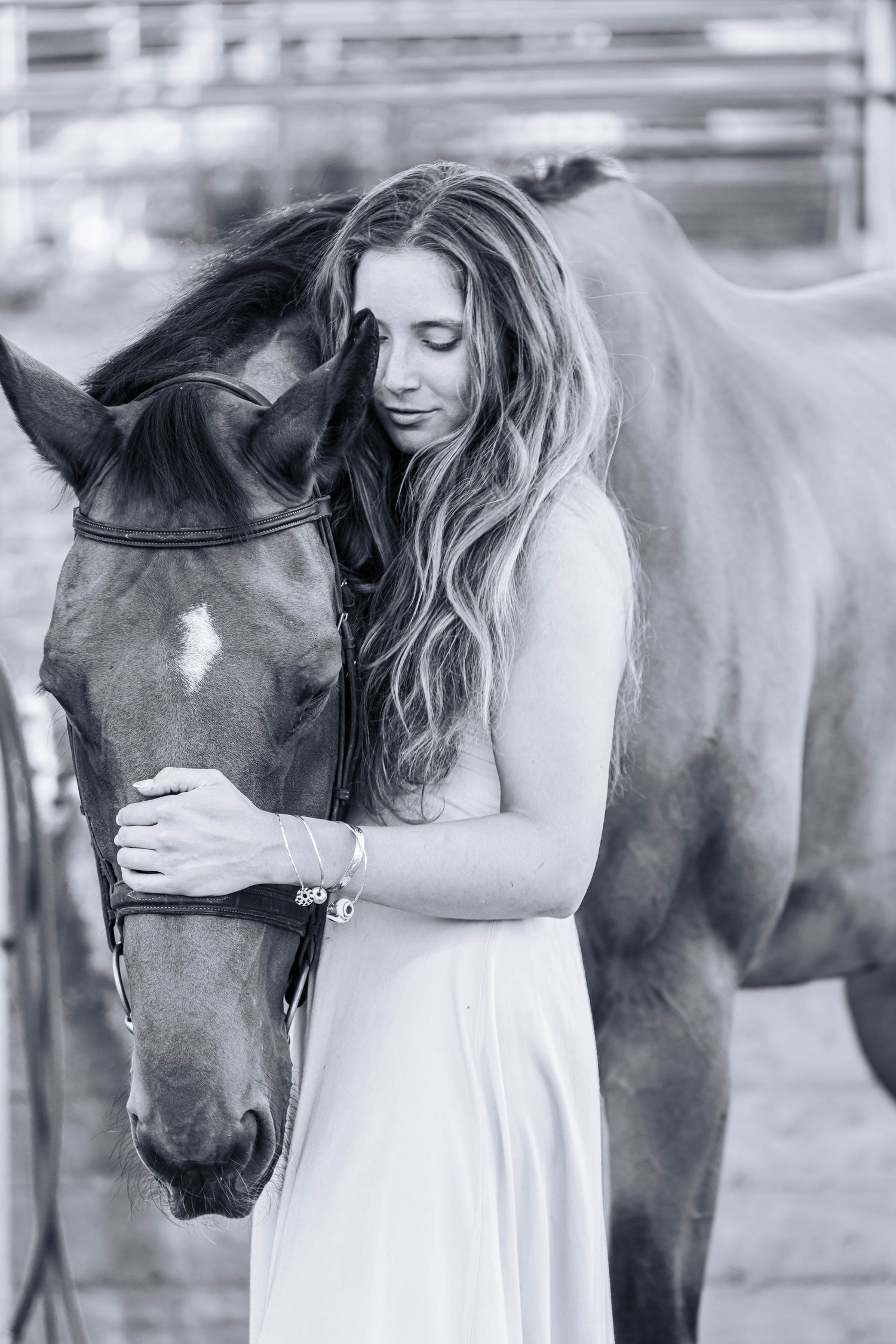 Black and white photo of a young woman in a white dress tenderly embracing a horse at a stable.