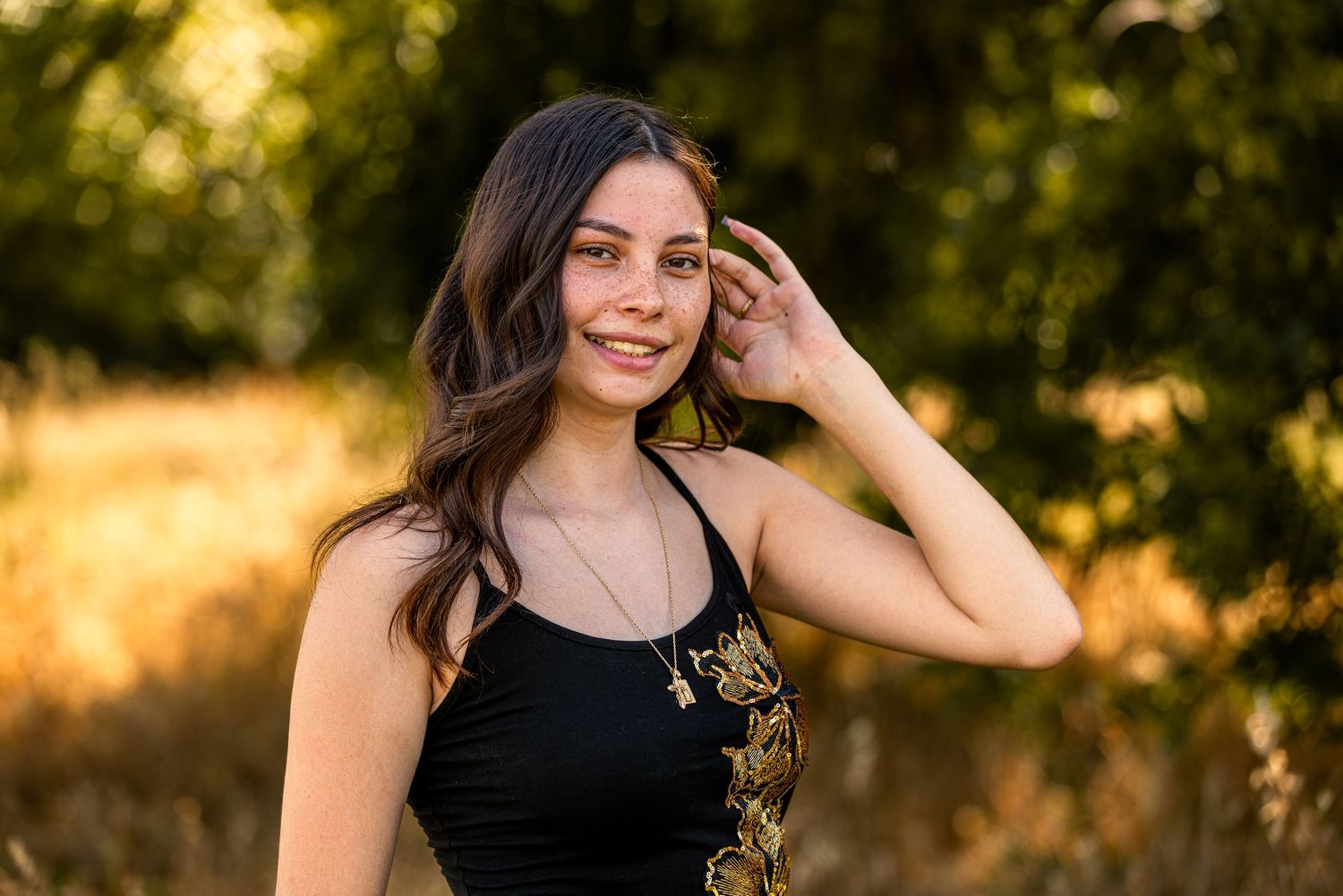 Young woman in black top smiling outdoors, golden sunlight filtering through trees in background.