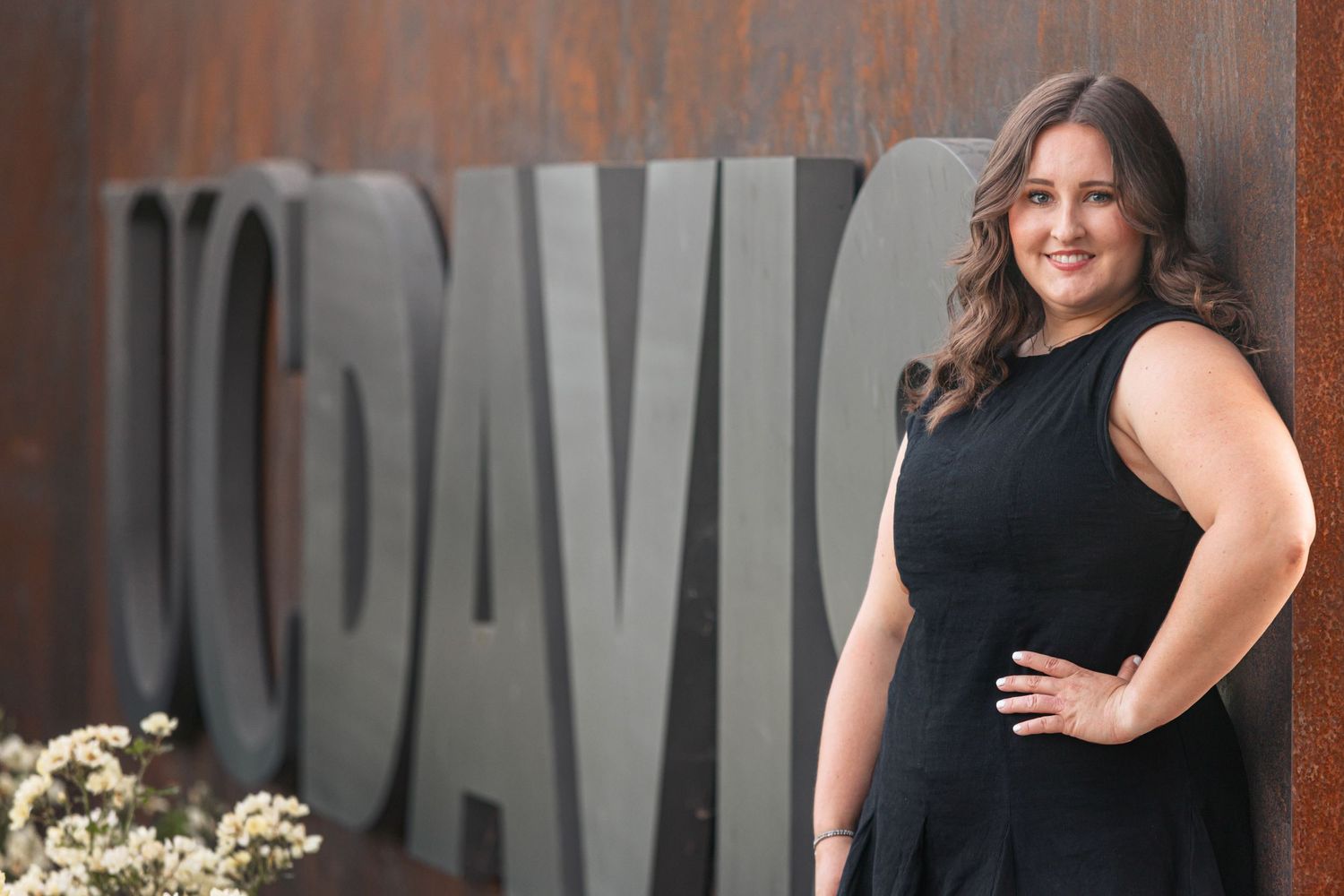 Smiling woman in black dress poses beside UC Davis metal lettering sign outdoors.