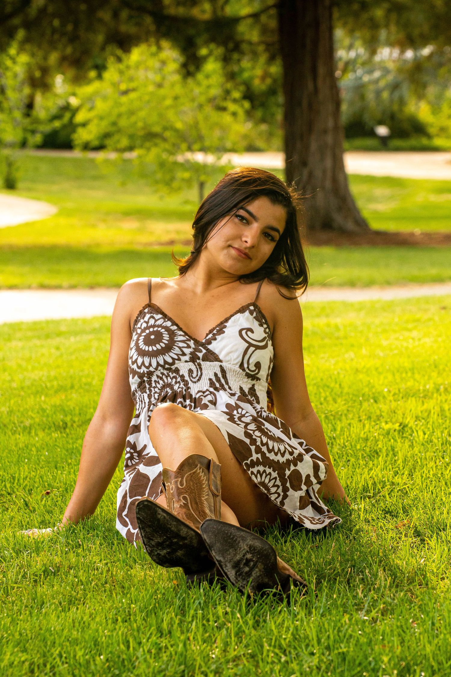Young woman in floral dress and cowboy boots sitting on green grass under a tree.