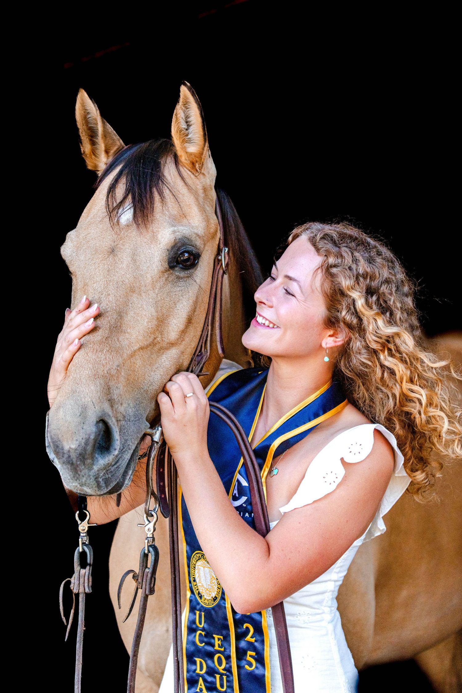 Smiling graduate in cap and gown sash posing with a tan horse in a dark stable.