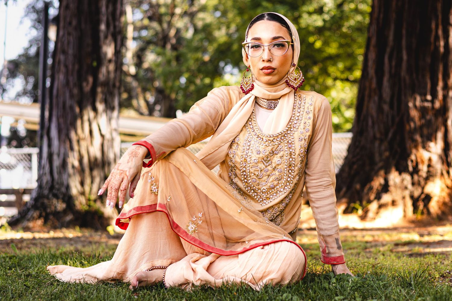 Woman in elegant gold Indian salwar kameez with dupatta sitting gracefully on grass near large trees.