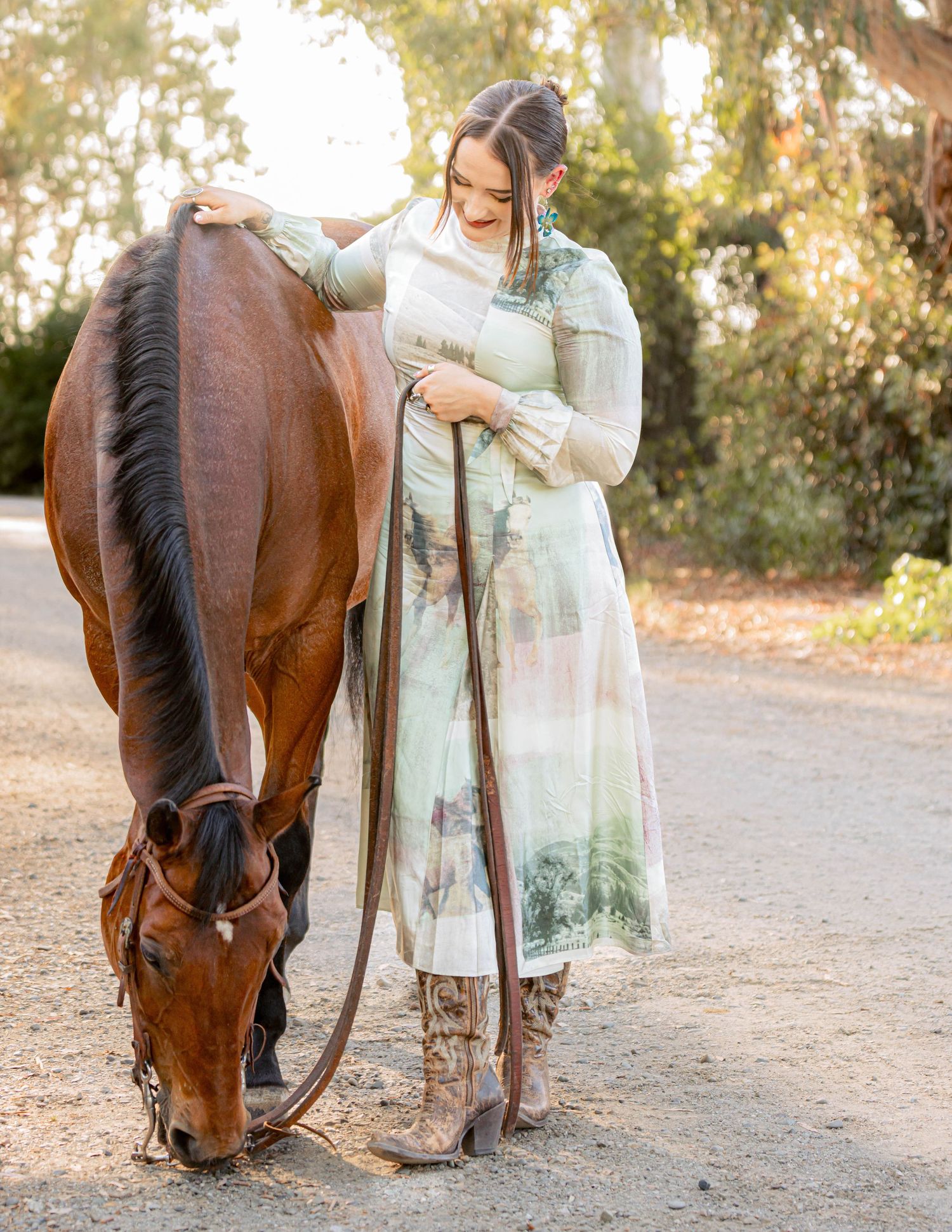 Woman in scenic print dress and boots stroking a grazing bay horse outdoors.