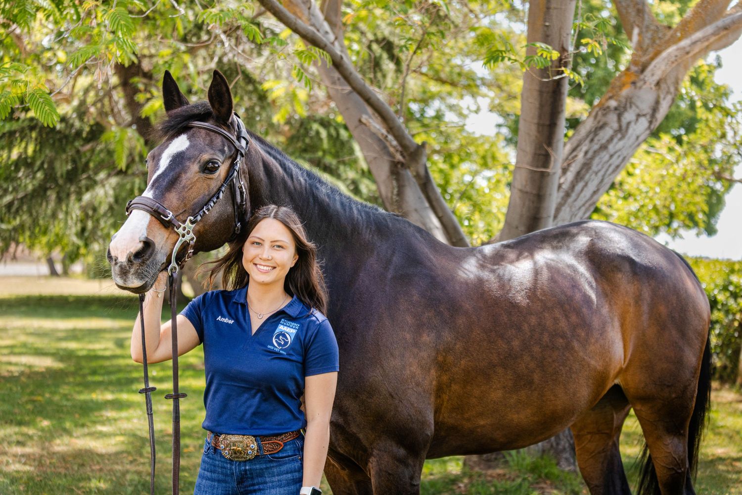 Young woman in blue polo smiling beside a dark bay horse in a green outdoor setting.