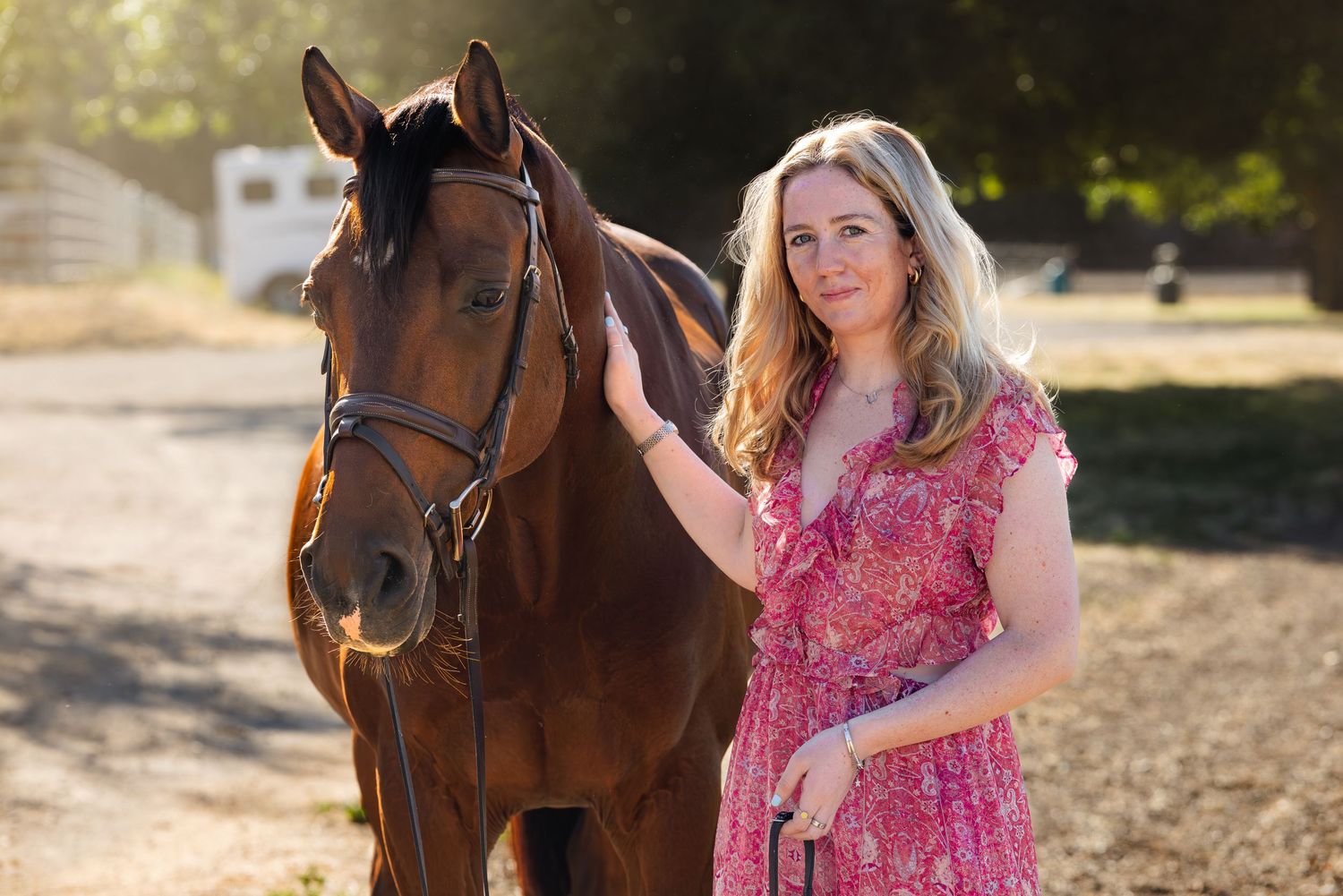 Woman in pink floral dress petting a brown horse outdoors in warm golden sunlight.