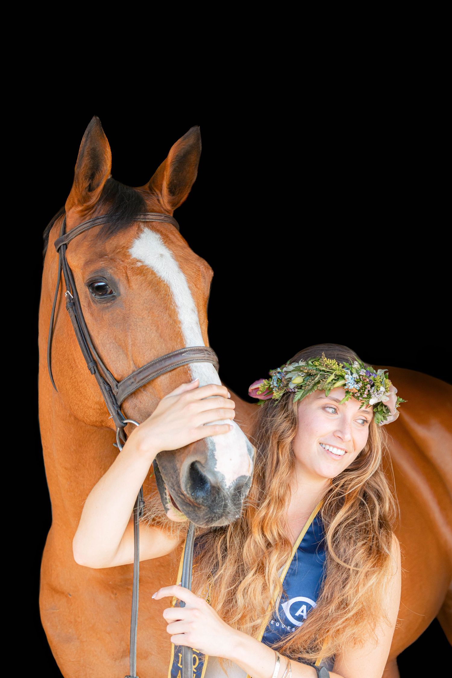 Smiling woman wearing a floral crown holds a bay horse by the bridle against a black background.