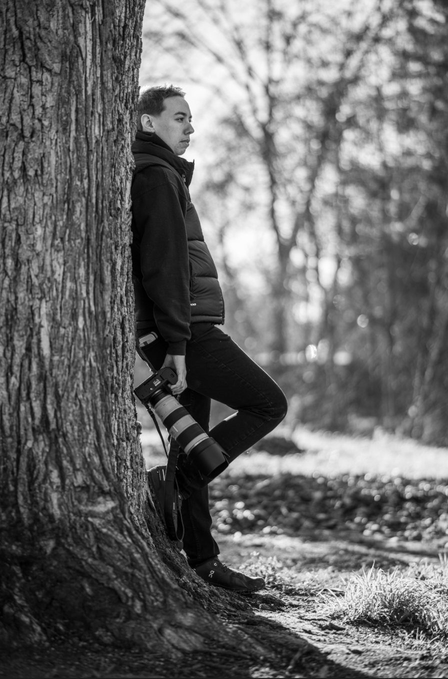 Black and white photo of young man leaning against tree trunk holding camera in winter forest.