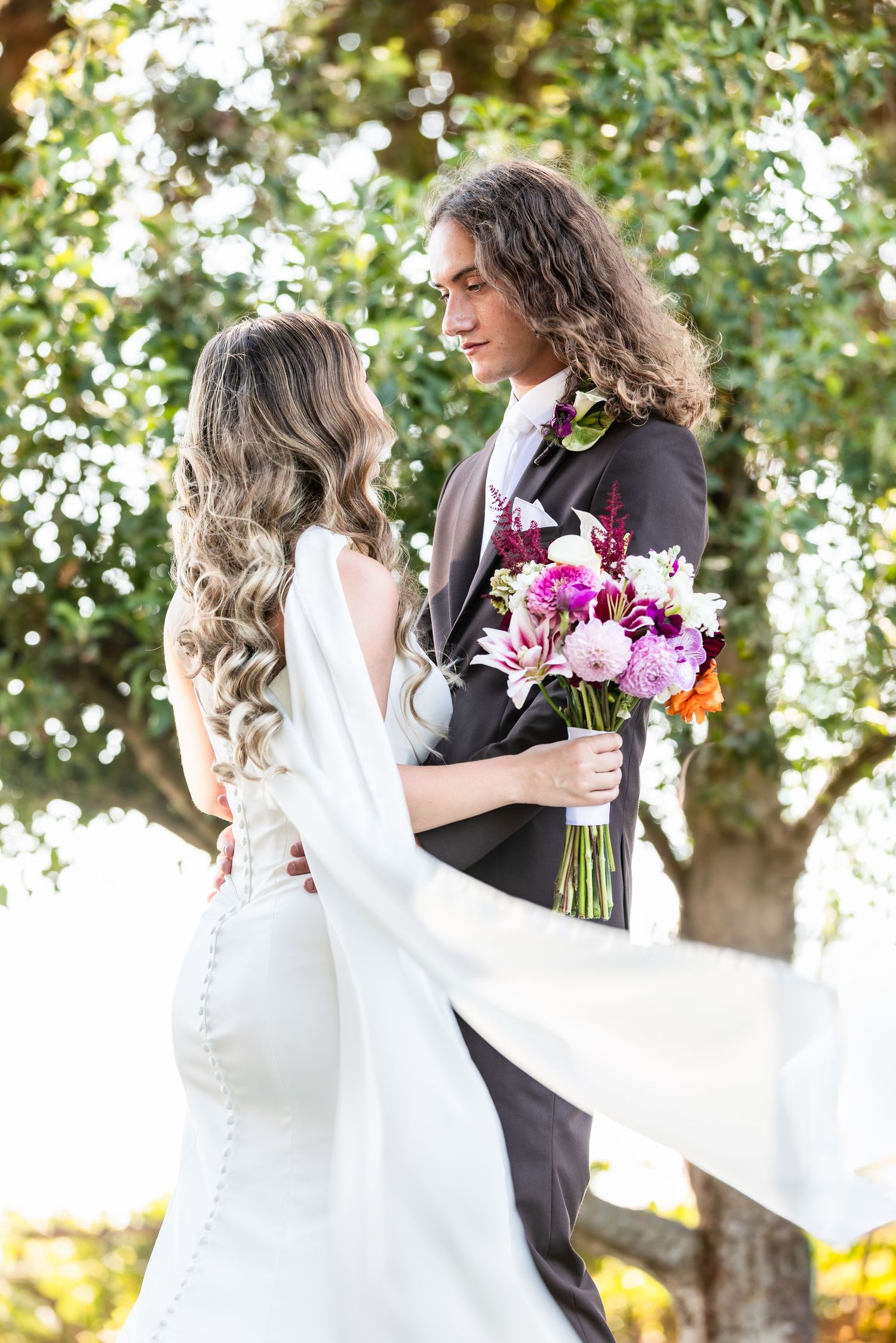 Bride and groom exchanging vows outdoors, holding a vibrant floral bouquet, with lush green trees in the background.
