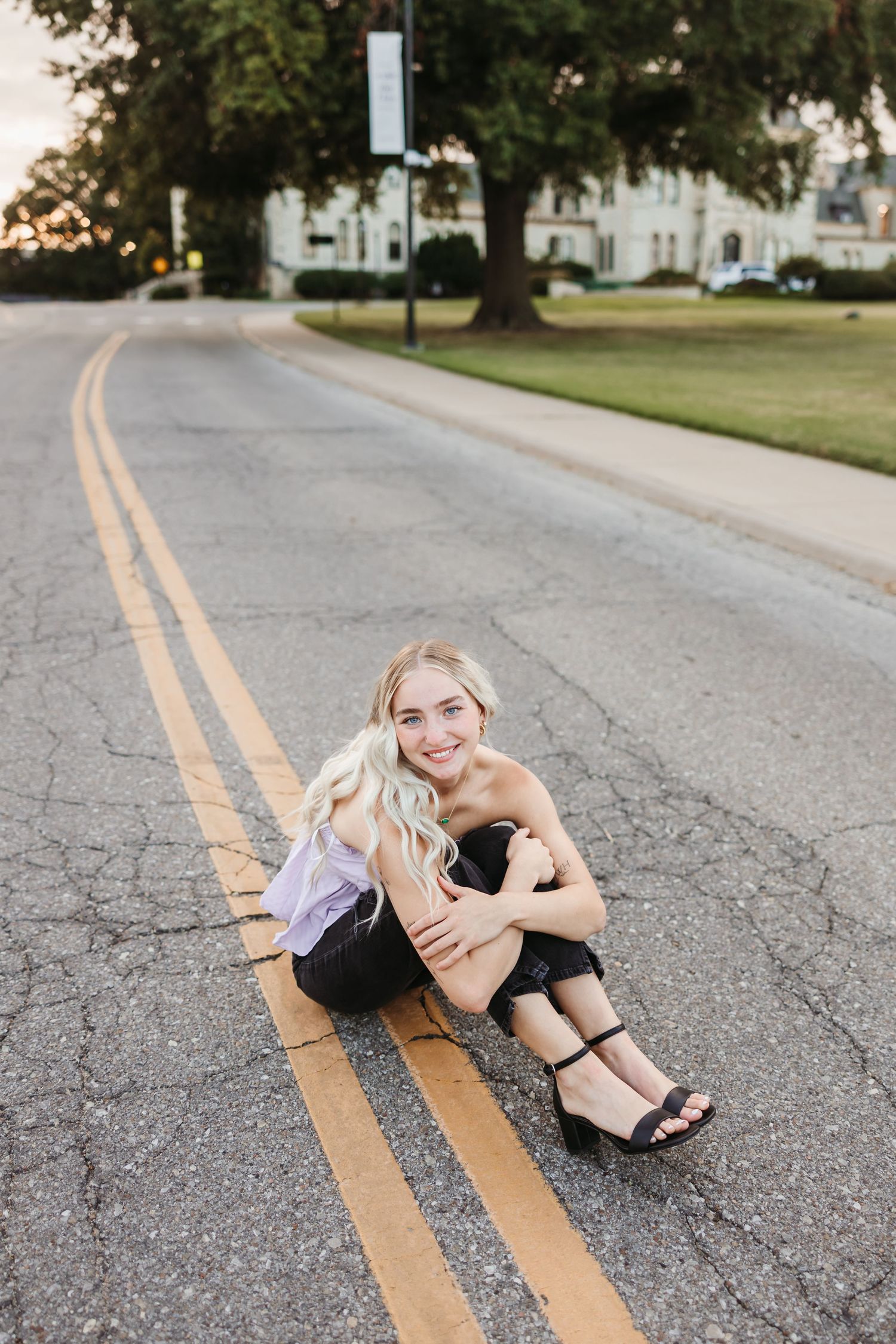 Smiling blonde woman sitting on road center lines in black outfit and heels, with tree-lined street and building in background.