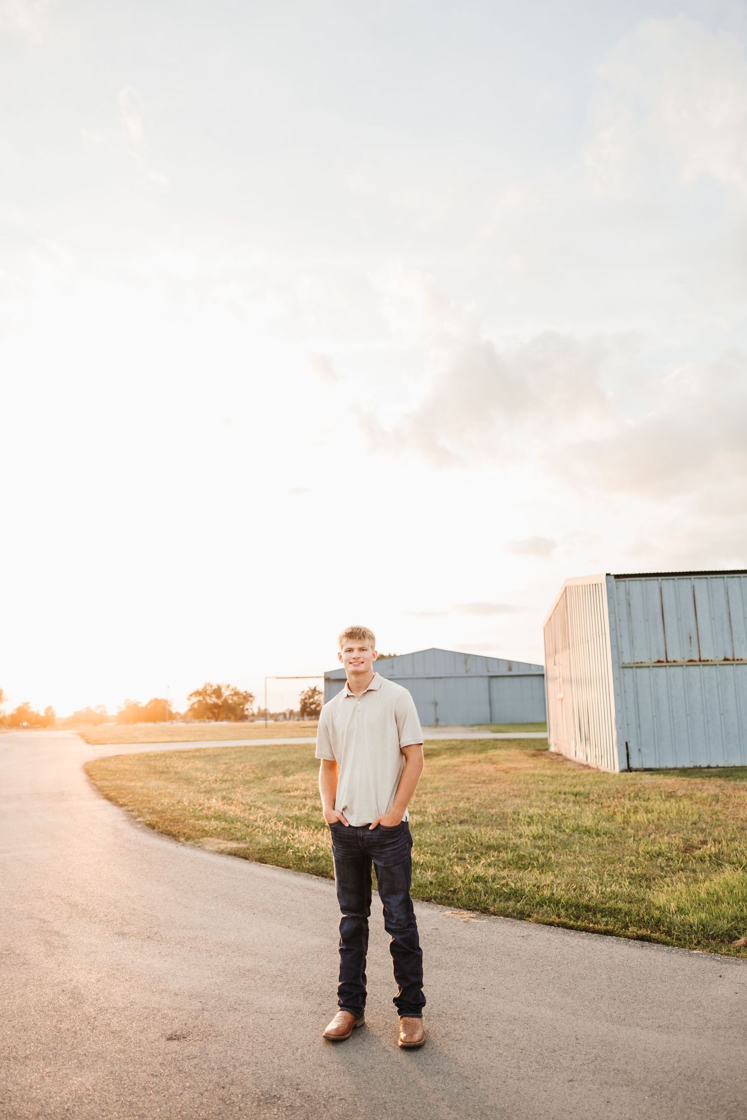 Young man in white polo and dark jeans stands on a rural road at golden hour near metal farm buildings.