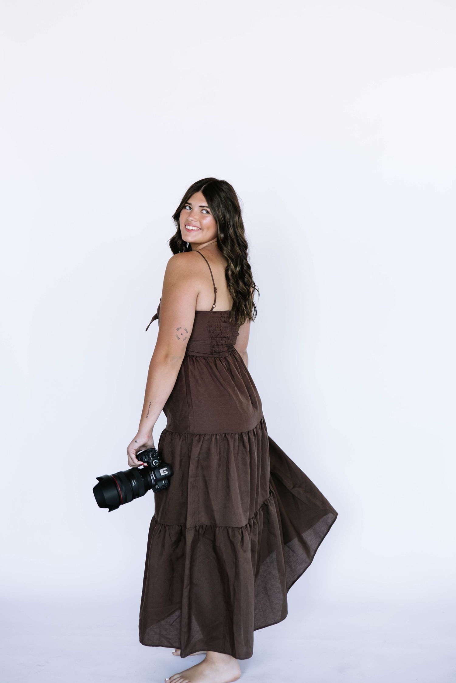 Photographer in brown maxi dress holding camera against white background.