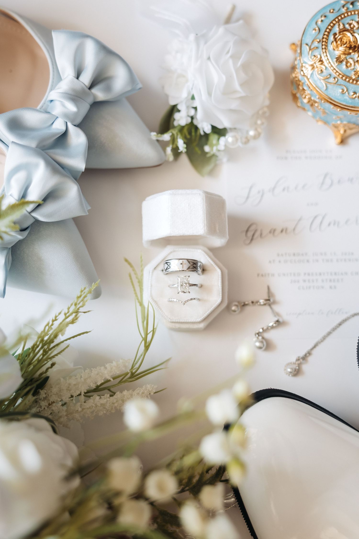 Elegant wedding flat lay with diamond rings in white box, blue bow heels, floral boutonniere, pearl necklace, and invitation.