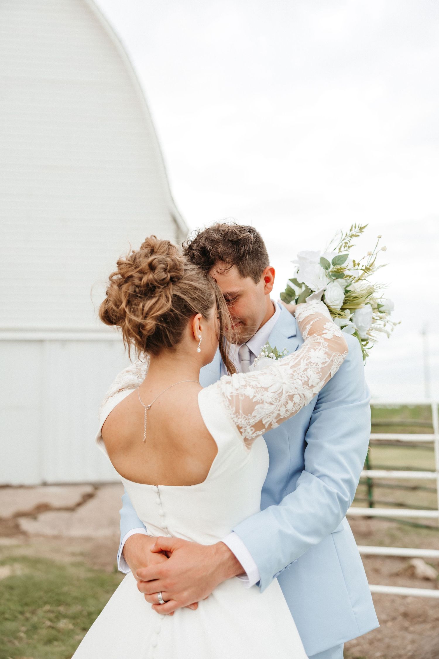 Bride and groom embracing outdoors near white barn, groom in light blue suit holding white floral bouquet.