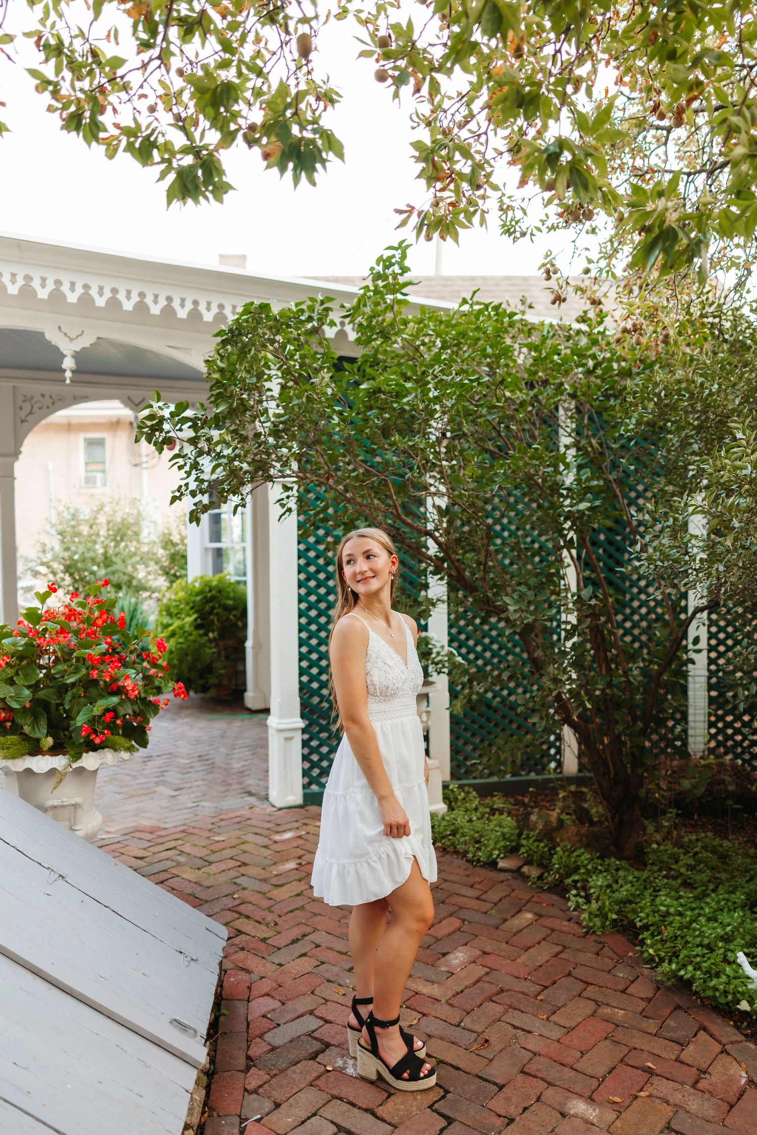Young woman in white summer dress and wedge sandals posing on brick pathway surrounded by lush garden greenery.