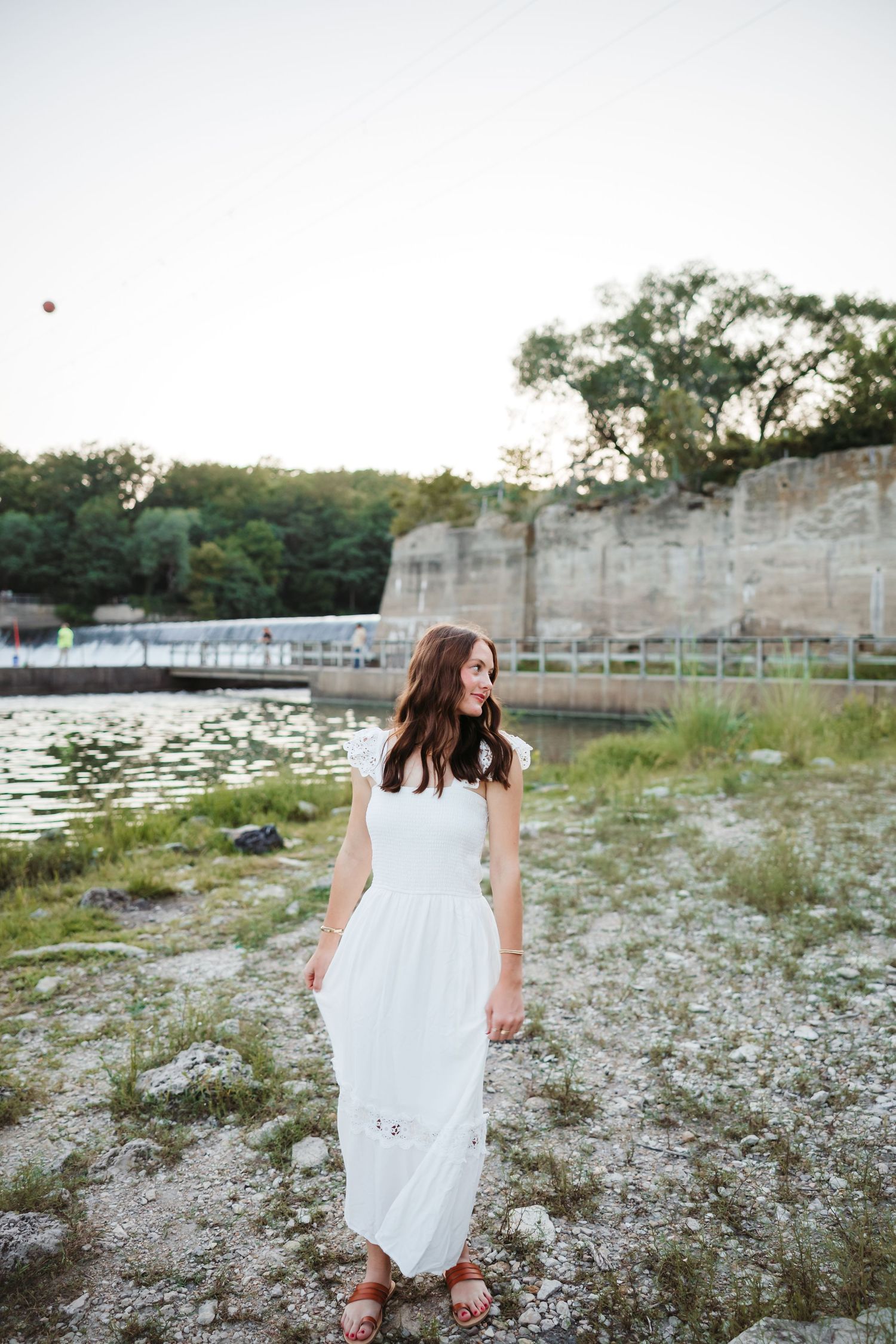 Person in white dress walking along rocky shoreline near stone wall.