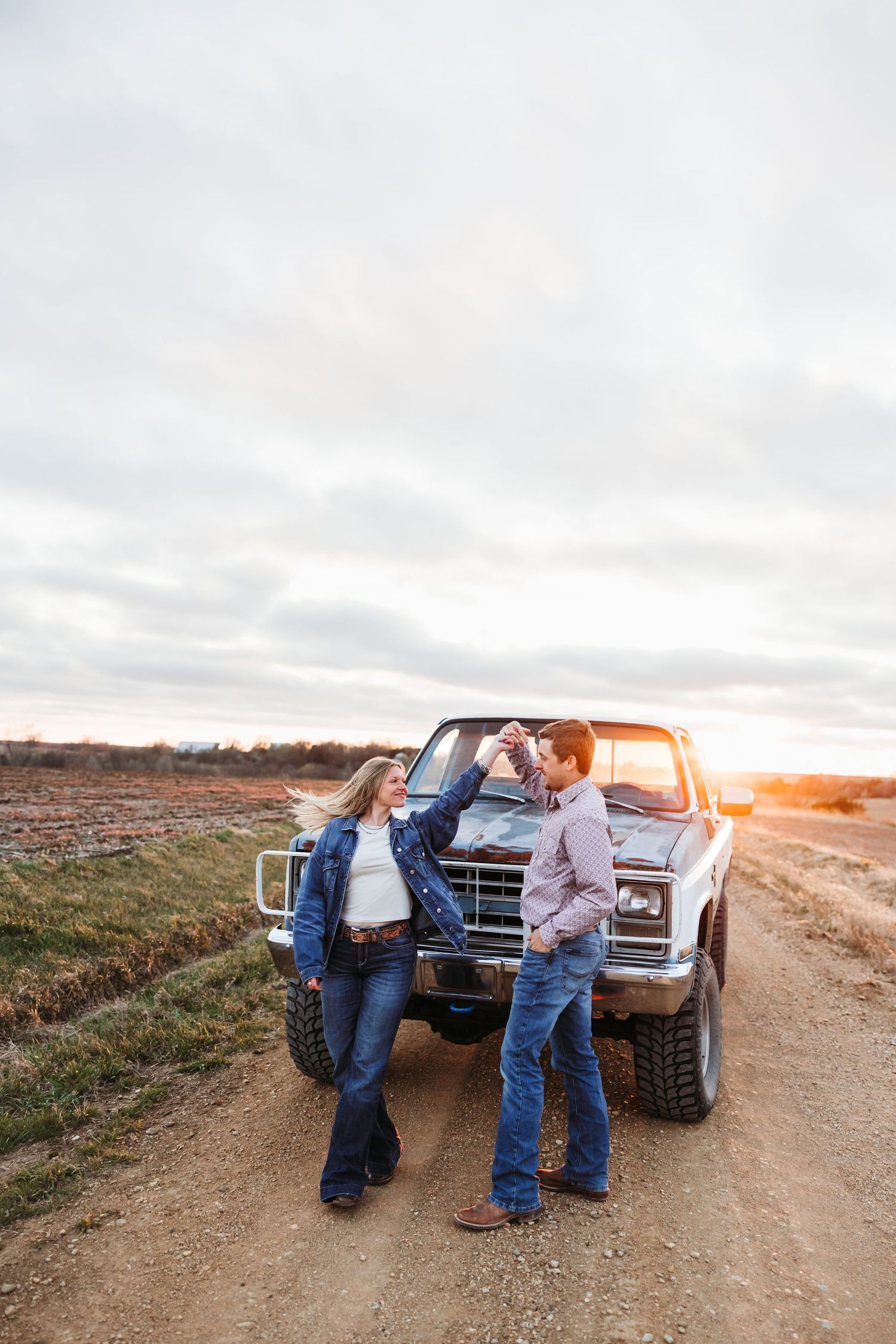 Couple dancing on a dirt road at sunset beside a classic truck in a rural field, both wearing denim.