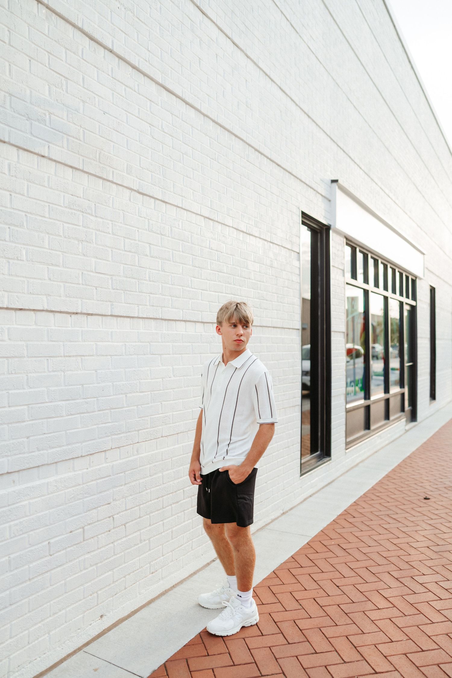 Young man in white striped shirt and black shorts posing against a white brick wall outdoors.