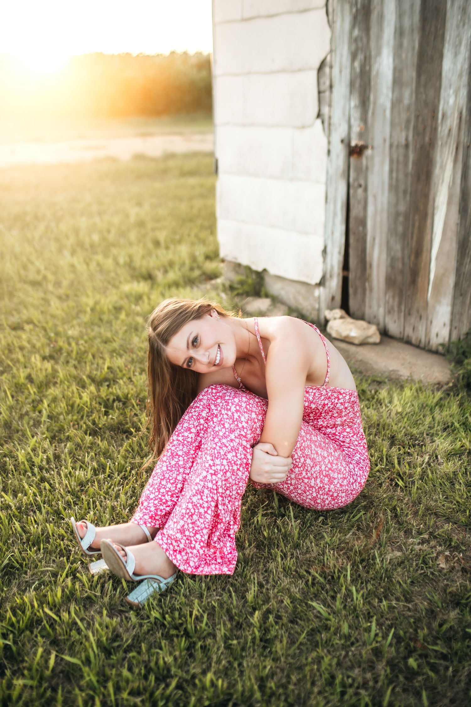 Young woman in pink floral jumpsuit sitting on grass near rustic barn door in golden sunset light.