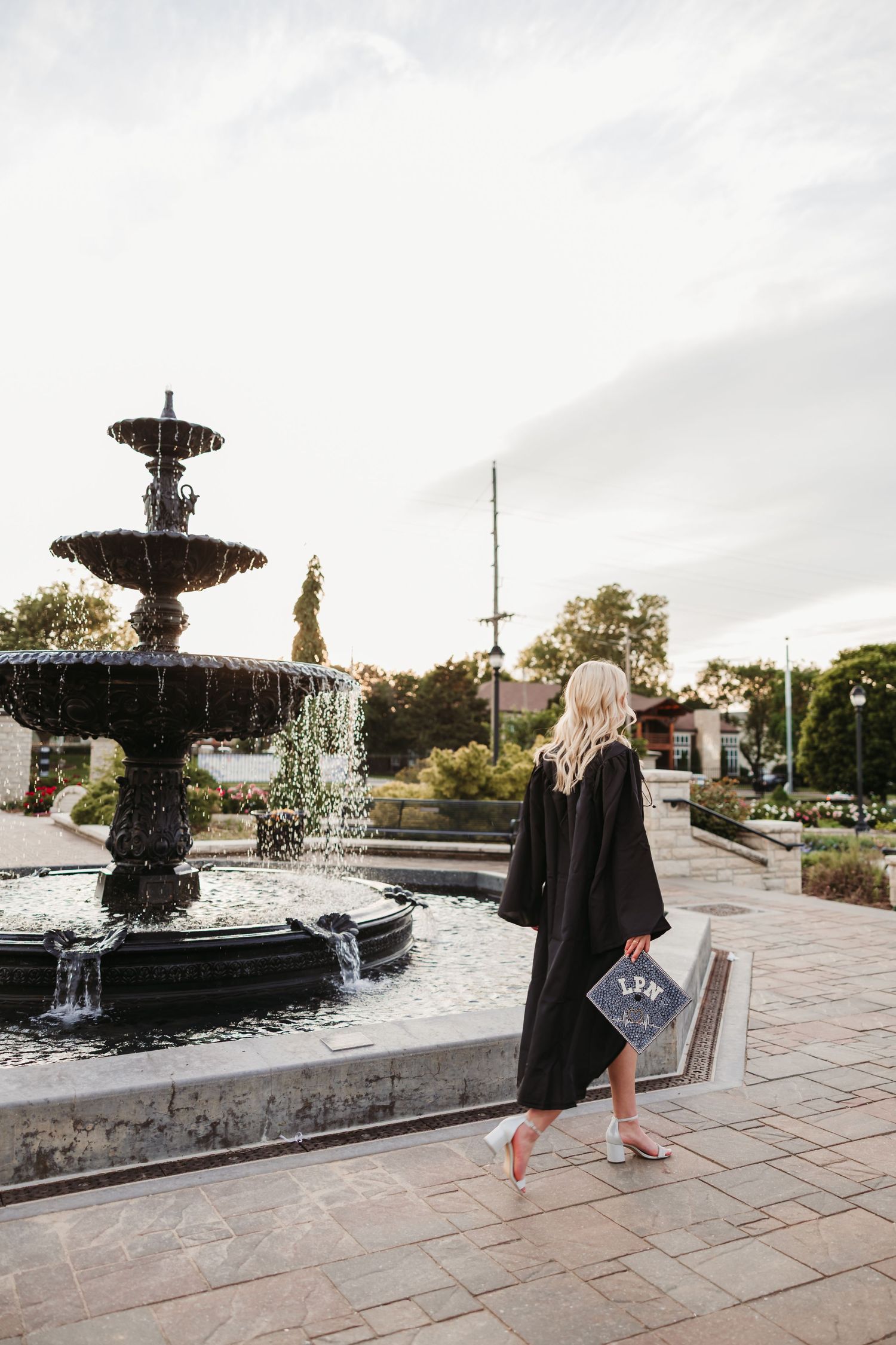 A blonde graduate in black gown holds decorated cap near ornate tiered fountain outdoors.