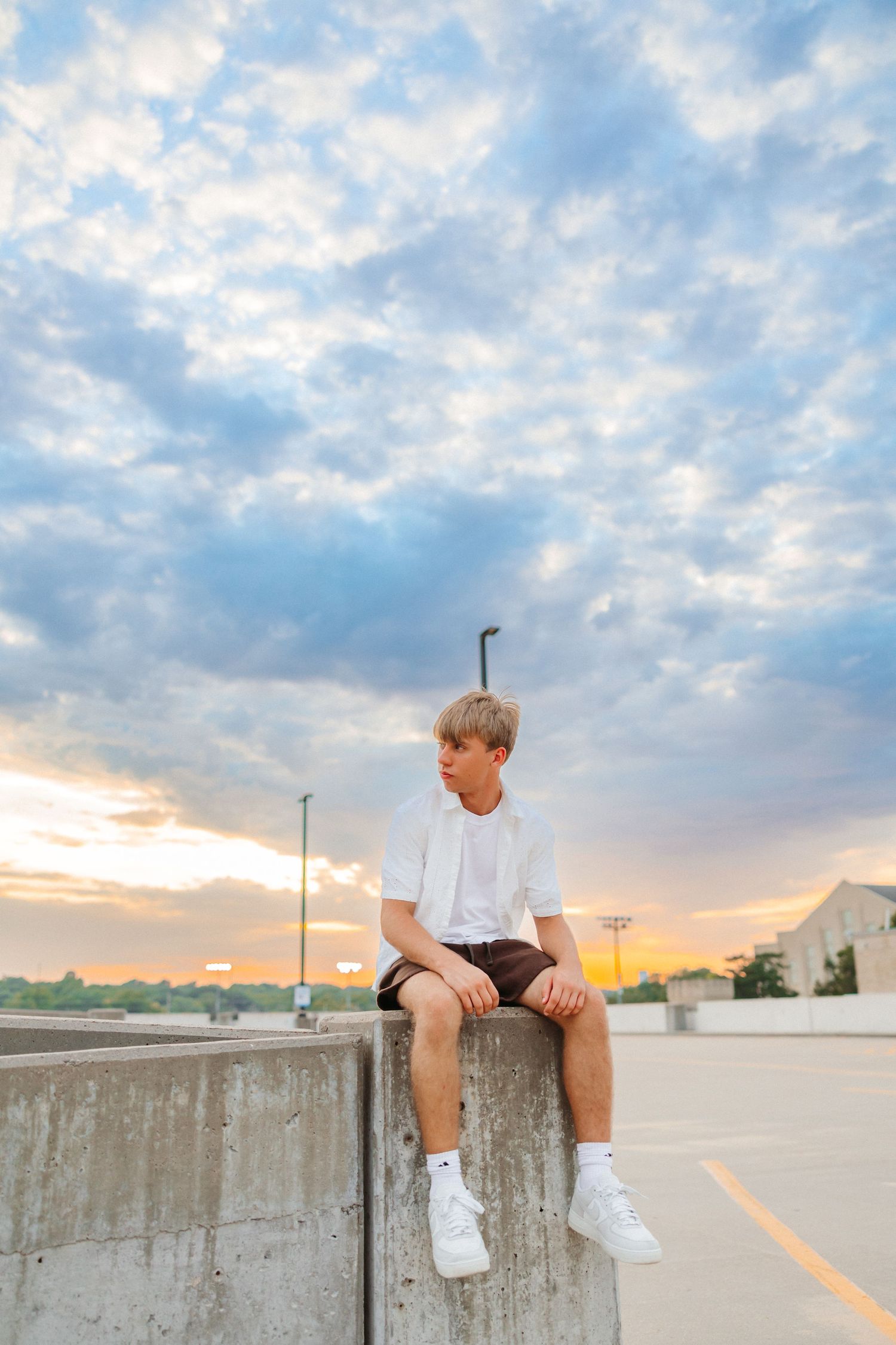 Teen boy in white tee and shorts sits on concrete wall at sunset under dramatic cloudy sky.