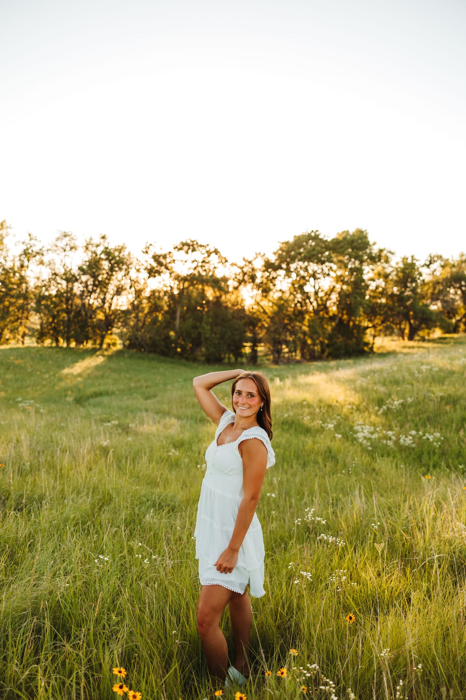 Young woman in white dress standing in sunny green meadow with wildflowers and trees at golden hour.